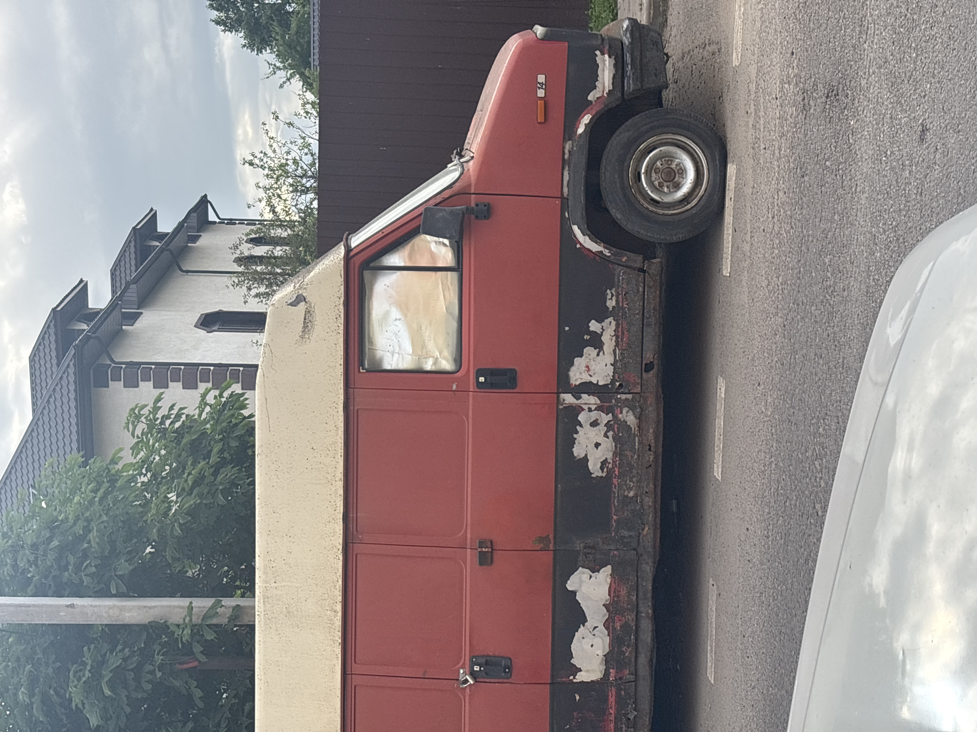 The image shows the side of a red and black panel van parked on a street in Misaylovo, Russia. The van appears to be old and worn, with paint peeling off the black sections. It has a cream-colored boxy extension on the back. The front of the van, including the driver's side window and a side mirror, is visible. The vehicle is parked next to a sidewalk and a road with white lane markings. In the background, a building with a tiled roof and some trees can be seen. The sky is cloudy. There is no activity or event taking place, and no people are visible in the image. A number "14" is visible on the front of the van. The overall impression is of a quiet, overcast day in a residential or semi-industrial area.