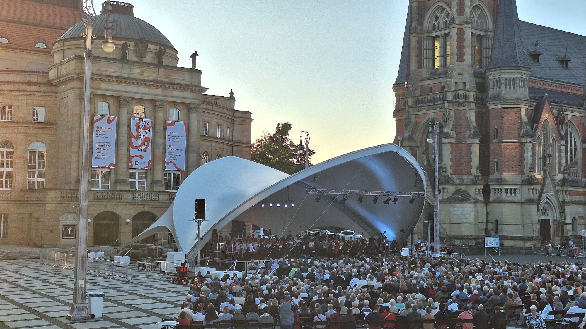 The image depicts an outdoor concert taking place in front of the Chemnitz Opera House and the adjacent Museum Gunzenhauser in Chemnitz, Germany. A large, white, arched tent covers a stage where an orchestra is performing. The audience, composed of a diverse crowd of people, is seated in chairs in front of the stage, facing the performers. The buildings in the background are classic examples of European architecture, with ornate detailing and grand facades. The sky is clear and a gentle sunset light bathes the scene, casting long shadows across the plaza. Several banners are visible on the side of the Opera House, advertising upcoming events. The overall atmosphere is one of cultural celebration and community gathering.