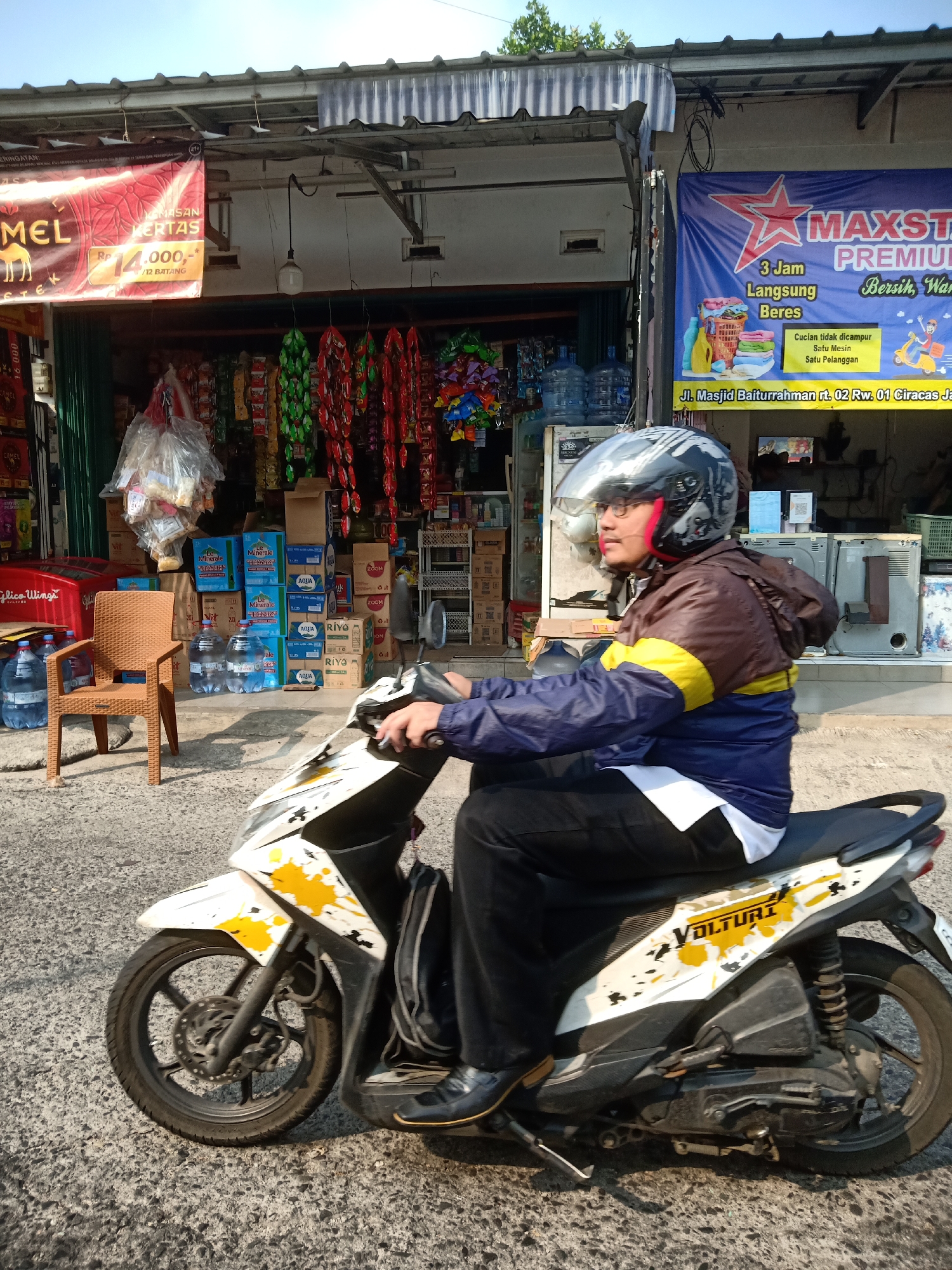 The image shows a man riding a white and yellow scooter down a paved street. The man is wearing a blue and brown jacket, black pants, and a black helmet with a silver visor. He is looking to his left. The scooter has a white body with yellow and black accents. In the background, there is a storefront with a blue banner that reads "MAXSTAR PREMIUM". To the left of the storefront, there is a red cooler and a brown plastic chair. Several large water bottles are stacked in front of the storefront. The scene appears to be in a commercial area. The banner in the background provides a clue to the location. The banner reads "Jl. Masjid Baiturrahman rt. 02 Rw. 01 Ciracas Jakarta Timur." This indicates the location is on Masjid Baiturrahman street, neighborhood 02, residential area 01, in Ciracas, East Jakarta, Indonesia.