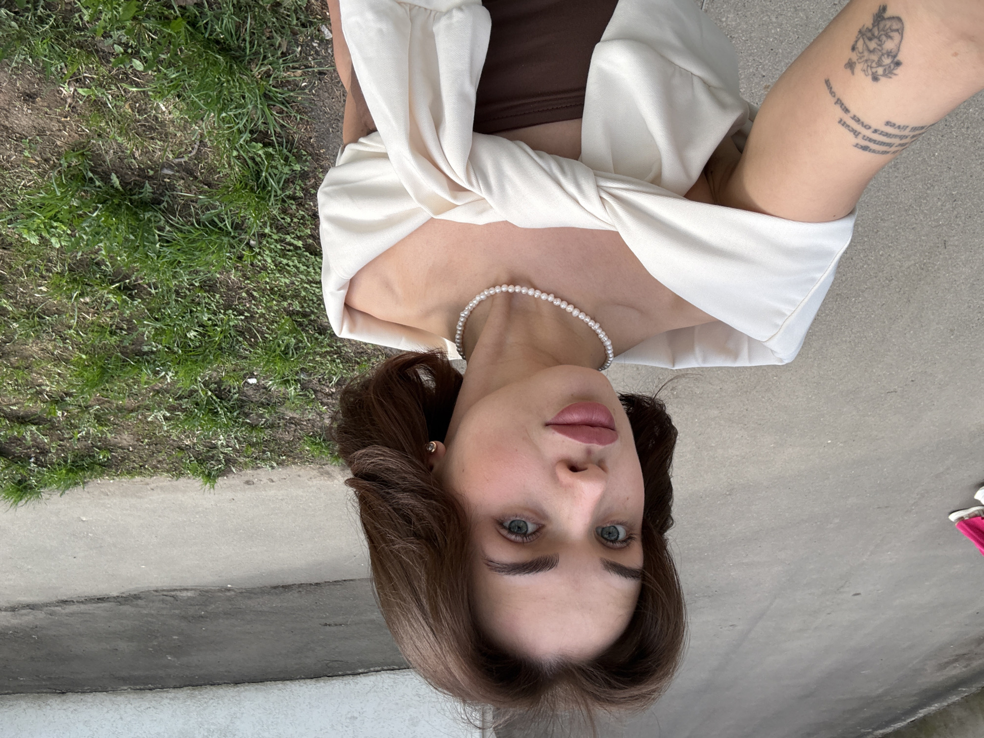 A woman is holding a phone above her head, photographing herself at a low angle. She has light skin, brown hair, and blue eyes. She is wearing a brown top, a white blouse, and a pearl necklace. There is a tattoo on her left forearm. She is standing outside on a paved area with grass in the background. The exact location in Sarapul, Russia is not discernible from the image.