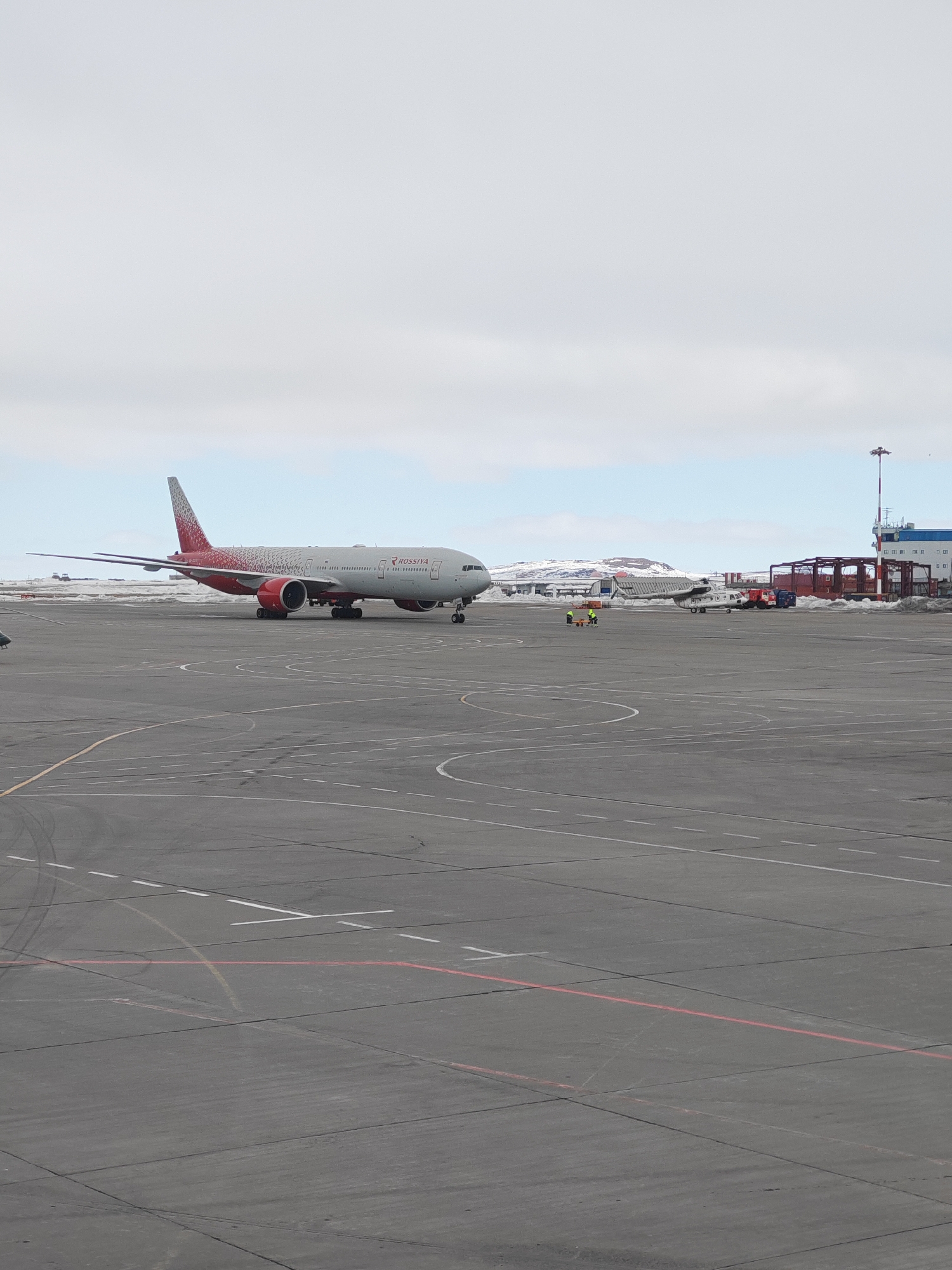 The image depicts an airport scene in Ugolnye Kopi, Russia. A large passenger airplane, identified as a Rossiya Airlines Boeing 777 due to the markings on its fuselage, is positioned on the tarmac. The plane is primarily white with red accents on the tail and engine housings. Further back, near some buildings and a control tower with red and white stripes, is a helicopter. Two individuals in yellow vests are standing on the tarmac. The sky is overcast, and there are snow-covered mountains in the background. The tarmac is gray and marked with white and red lines.