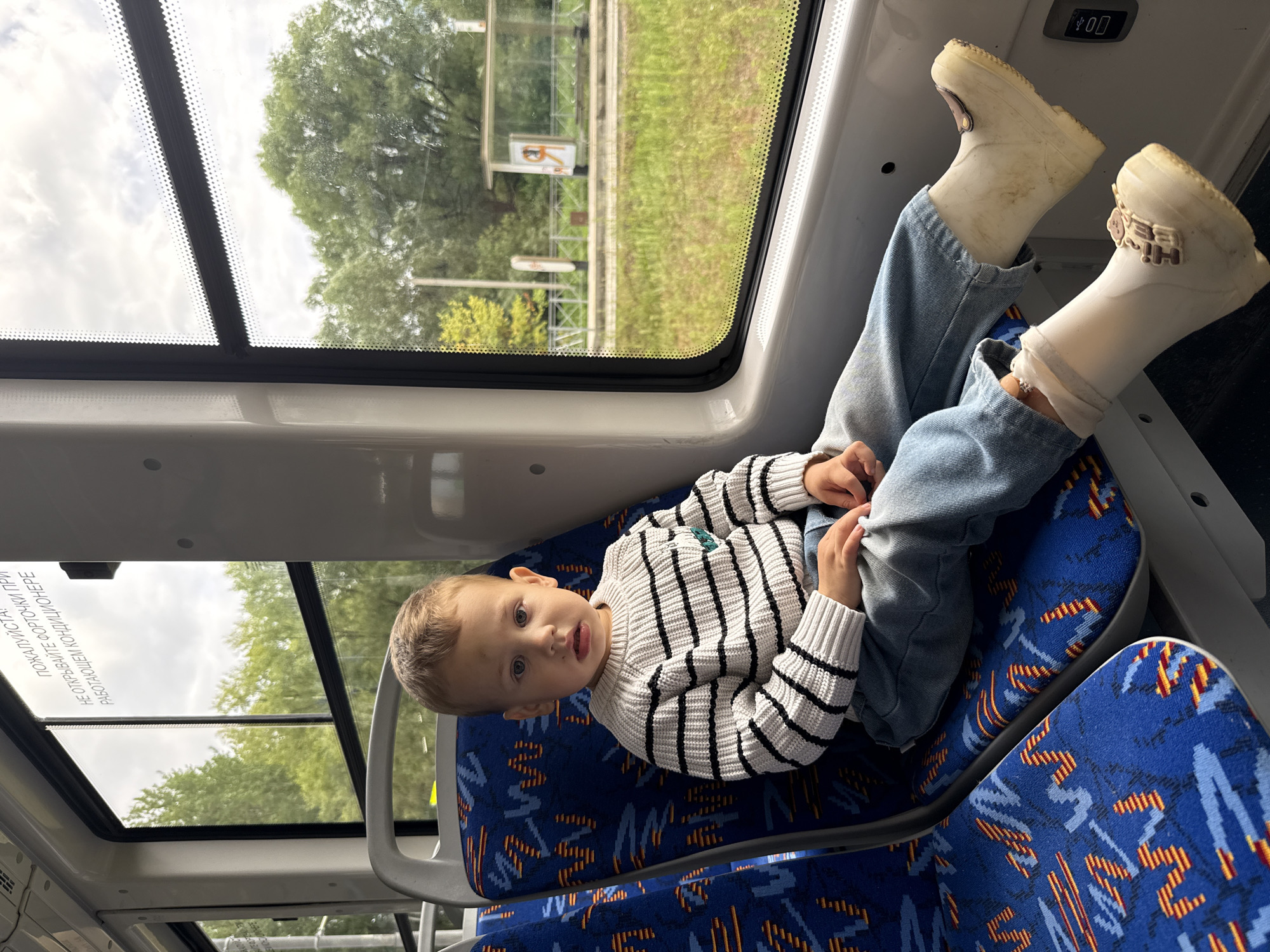 A young boy sits on a blue and orange patterned seat inside a public transport vehicle, likely a bus or train, with a window to his right. He is wearing a white sweater with black horizontal stripes, light blue jeans, and white rubber boots. His gaze is directed slightly upwards and to his left. Outside the window, green trees and a cloudy sky are visible, with a bus stop shelter in the distance. The text on the window, "ПОЖАЛУЙСТА! НЕ ОТКРЫВАЙТЕ ФОРТОЧКИ ПРИ РАБОТАЮЩЕМ КОНДИЦИОНЕРЕ" translates to "PLEASE! DO NOT OPEN WINDOWS WHEN THE AIR CONDITIONER IS ON." This indicates the location is Rusanovo, Russia. The interior of the vehicle features grey plastic seating and white paneling. The lighting suggests it is daytime.