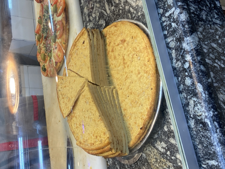 A round, light-yellow frittata or tortilla on a silver tray has been partially cut into slices. Some slices are grouped together, while two triangular pieces sit beside them. In the background, a pizza with tomatoes and green olives is visible. A granite countertop is partially visible on the right, and a reflective glass surface covers the front.