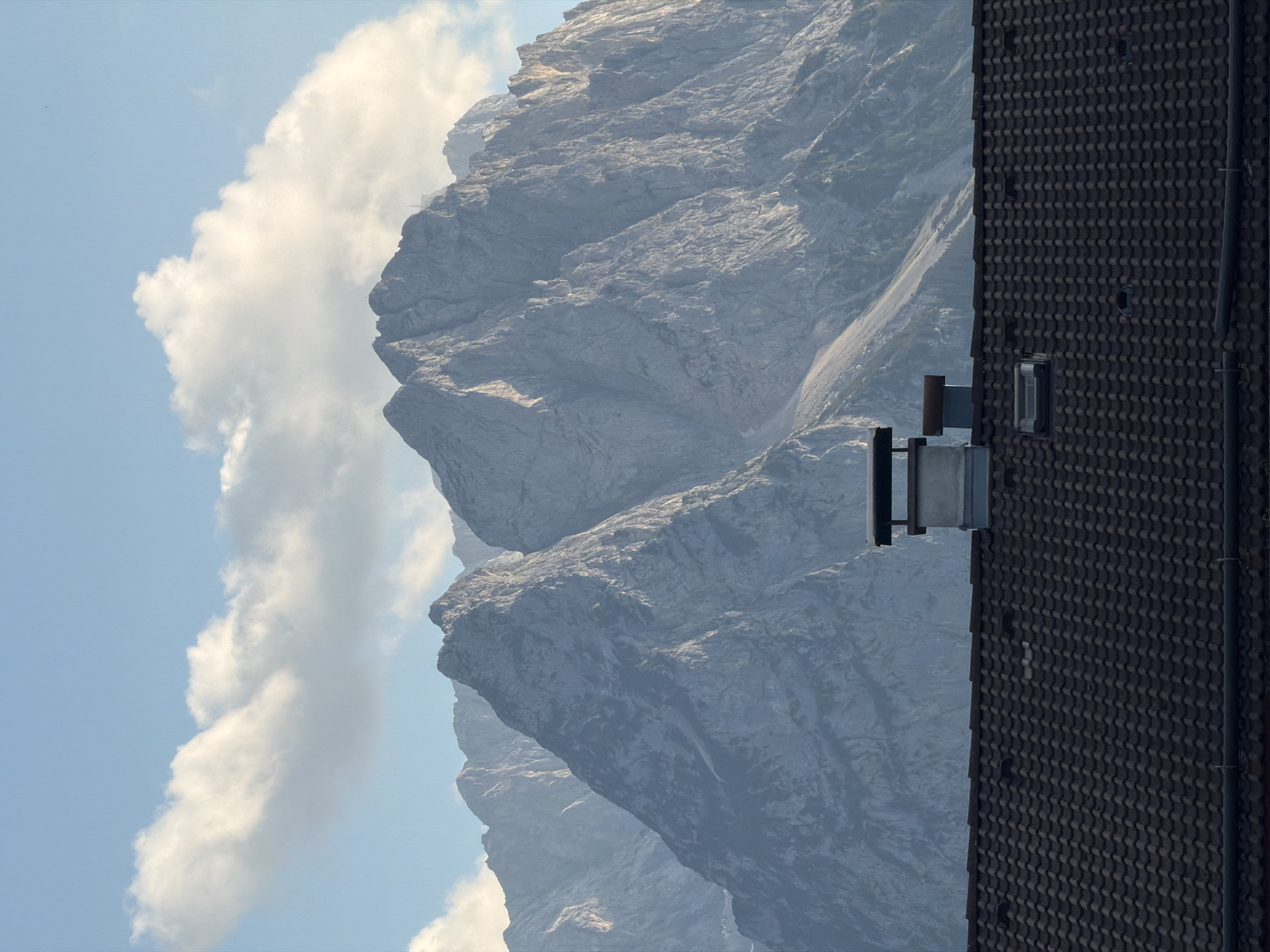 The image shows a rooftop in the foreground with a dark brown, tiled surface. A chimney with metal caps is visible on the roof, along with a small skylight. Behind the rooftop, a majestic mountain range dominates the scene, its peaks jagged and rocky. The mountains appear to be covered in patches of snow and scree, with steep slopes and dramatic rock formations. The sky above is a clear blue, with a few wispy white clouds scattered across it. The lighting suggests it is late afternoon or early evening, as the sun casts long shadows and highlights the textures of the mountains. The overall scene evokes a sense of natural beauty and grandeur, with a hint of human habitation in the foreground.