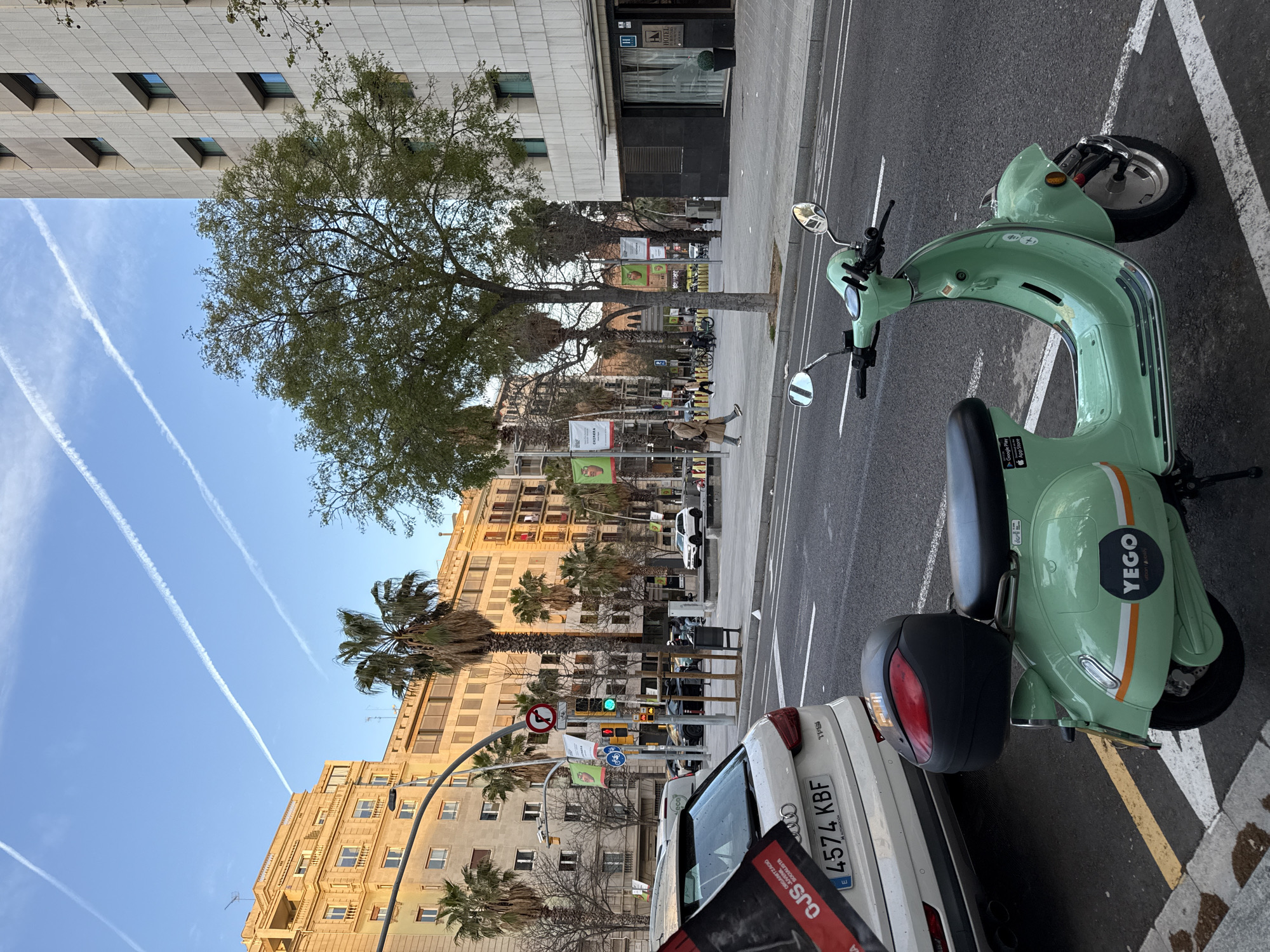The image depicts an urban street scene in Barcelona, Spain, specifically on Avinguda Diagonal, near the intersection with Passeig de Gràcia.
In the foreground, a mint green Yego-branded electric scooter with a black top case is parked at an angle on a designated parking spot marked with white lines on asphalt. Chrome rearview mirrors are visible. Behind the scooter, partially visible, is the rear of a white Audi Q3 TFSI SUV with the Spanish license plate "4574 KBF." A red and black banner, partially reading "OJS ORGANIZACIÓ JOVENTUT SOCIALISTA," hangs from the rear driver's side of the Audi.
The street extends into the midground with a traffic light showing a green signal, and a "No Turn" sign (red circle with a black arrow and a red slash) and a "Bicycle Lane" sign (blue circle with a white bicycle icon) are mounted on an overhead street lamp structure. Several large palm trees and deciduous trees line the wide sidewalk to the right. A male pedestrian in a light-colored coat walks right-to-left on the sidewalk in the middle ground, past a row of shared bicycle racks. Banners advertising "CHUSANA" are visible on lampposts along the avenue.
In the background, the left side features multi-story, classic Eixample-style buildings with yellow-orange facades, balconies, and numerous windows, illuminated by late afternoon sunlight. The right side of the background is dominated by a taller, modern building with a grey, textured facade and rectangular windows, identified as a hotel (Hotel Condes de Barcelona) with its entrance visible at ground level. The sky above is clear blue with three prominent white contrails from aircraft.