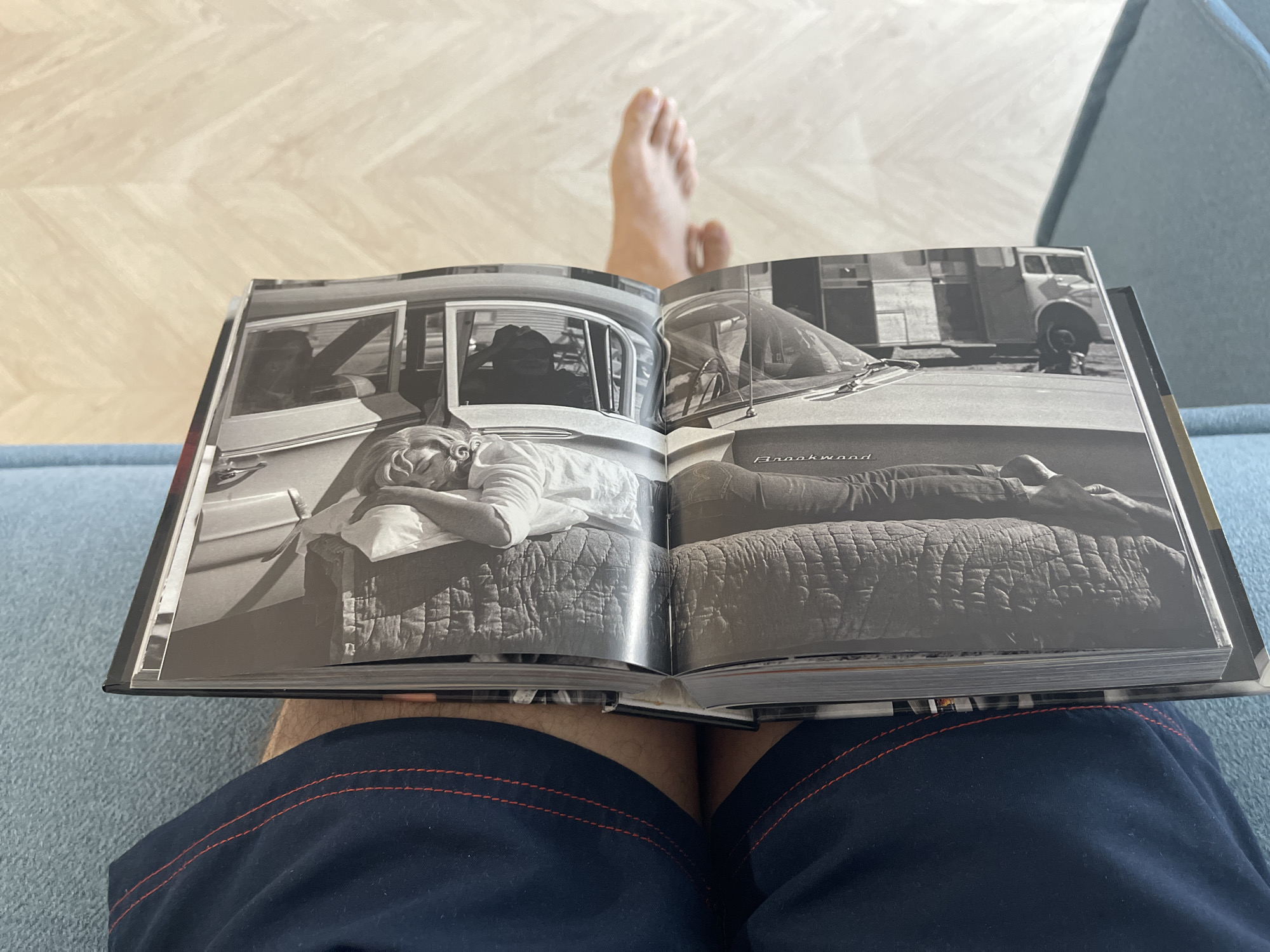 A photograph shows an open book resting on the lap of a person wearing blue shorts with red stitching. The open pages of the book display a black and white photograph of a man sleeping on a quilted bed inside a car. The car's window is open, and the side of a truck is visible in the background. A person's foot is visible in the upper part of the image. The person is seated on a blue couch in a room with a light-colored parquet floor.