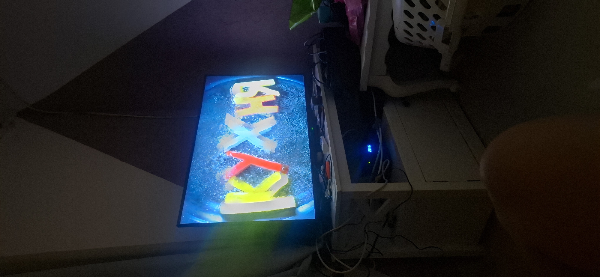 The image shows a dimly lit room with a large television screen displaying the word "КУХНЯ" in large, colorful, three-dimensional letters against a blue, watery background. The letters appear to be made of ice or a similar translucent material, with yellow, red, and white colors. Below the television is a white cabinet with various electronic devices and cables on top, including what appears to be a set-top box with a blue digital display showing "23:15". To the right of the cabinet is a white table with a black basket and other items on it. A white laundry basket is visible in the lower right corner. The room's walls have a patterned texture, with a dark triangular section on the right. The image is taken from a low angle, with a blurred object in the foreground, likely a person's leg or knee. The overall scene suggests a living room or entertainment area. No specific location in Yerocham, Israel is identifiable.