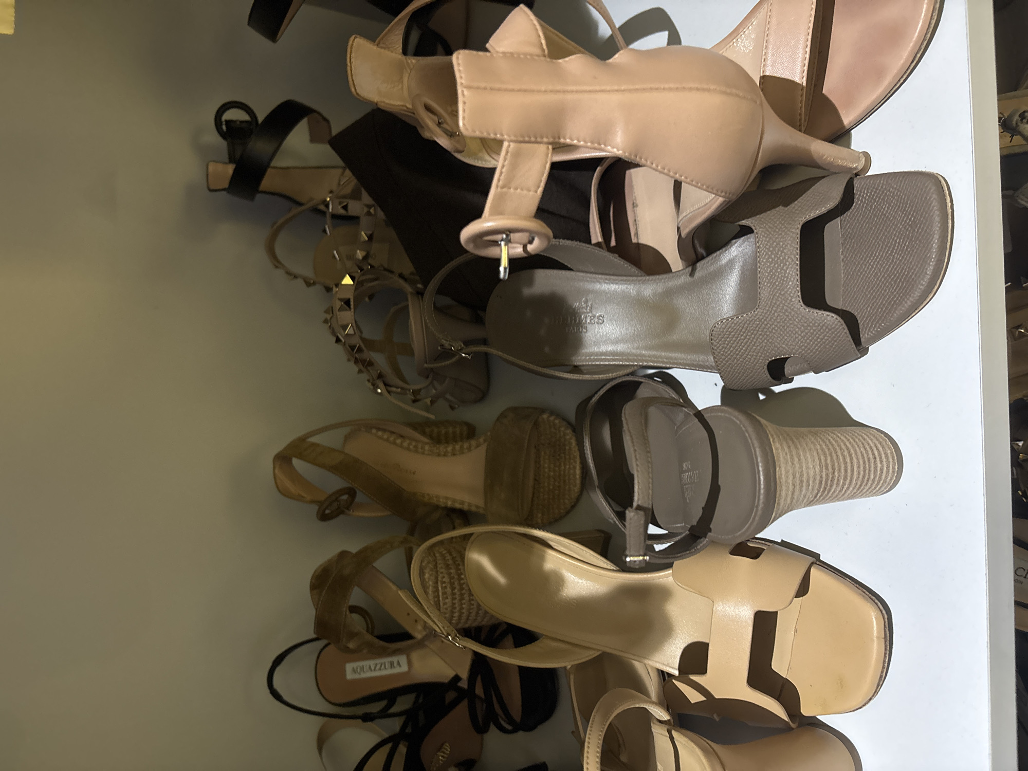 The image shows a collection of women's heeled sandals and shoes neatly arranged on a white shelf, likely in a closet or boutique. The shoes are of various designs and colors, including nude, tan, black, and brown. Some feature strappy designs, while others have block heels or pointed toes. A few pairs have embellishments like studs or woven details. The lighting suggests an indoor setting, possibly a retail environment or a personal collection display. The image does not provide any information that would allow for localization to Dubai, United Arab Emirates.
