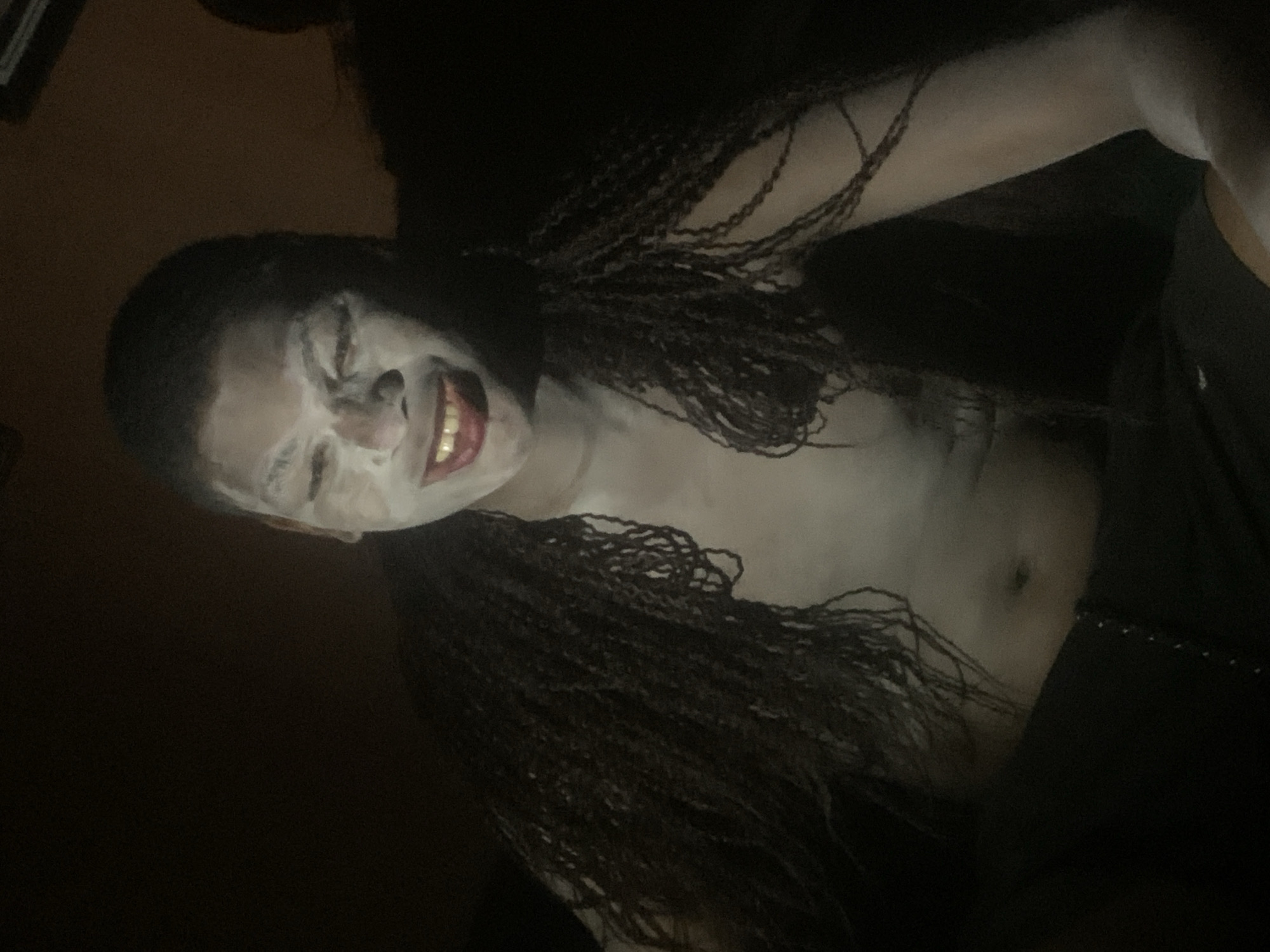 A person with braids lies tilted, facing upwards, with their face covered in white makeup and a red lipstick. They are smiling widely, exposing teeth. Their torso is bare and also covered in the white makeup. They are wearing dark clothing. The background is dark and mostly obscured. There is no visible indication of being in Onitsha, Nigeria.