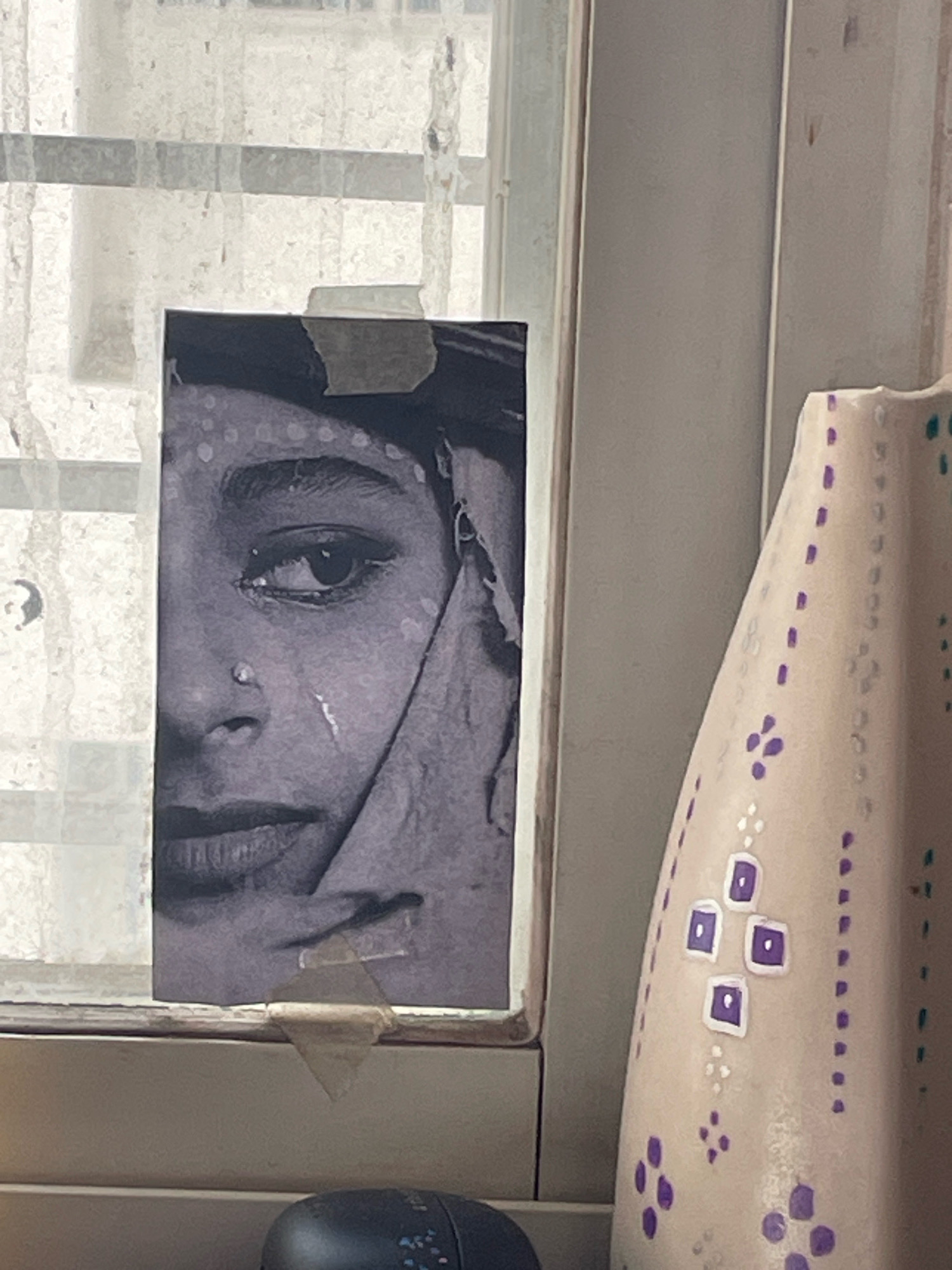 A close-up, medium shot captures a black and white photograph of a woman's face, partially taped to a window pane. The photograph is cropped to show her eye, nose, and lips, with a tear streak visible on her cheek. The window is covered with a sheer, textured curtain, obscuring any view of the outside environment. To the right of the photograph, a decorative ceramic vase with purple and white floral patterns stands on a surface. In the foreground, a dark, oval-shaped object, possibly a computer mouse, is visible. The scene appears to be indoors, near a window, in Loni Kalbhor, India.
