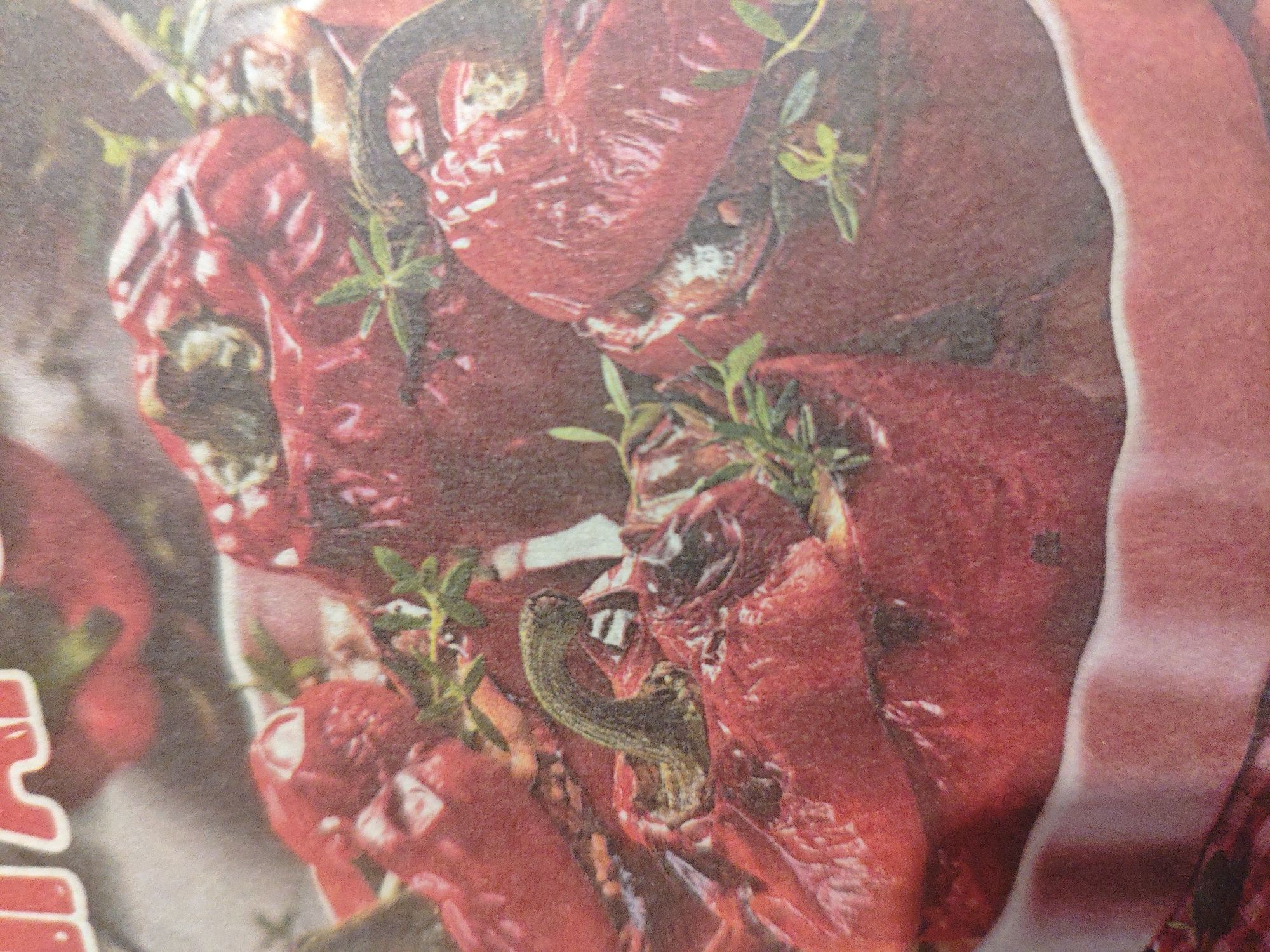 The image is a close-up shot of a portion of what appears to be a newspaper or magazine advertisement. The dominant subject is a cluster of bright red bell peppers, some appearing whole and others cut open. Interspersed among the peppers are sprigs of fresh green herbs, likely thyme or rosemary. A dark, elongated object, possibly a piece of dark chocolate or a sliver of dried fruit, is coiled on top of one of the peppers. The background is a reddish-brown hue, blending into the peppers themselves. On the left side of the frame, a segment of white-outlined, red block lettering is visible, though the text is incomplete and illegible. The overall impression is one of culinary presentation, possibly for a recipe or food product. The image does not contain any elements that would allow for specific identification of its location within Armavir, Russia, or any other geographical context.