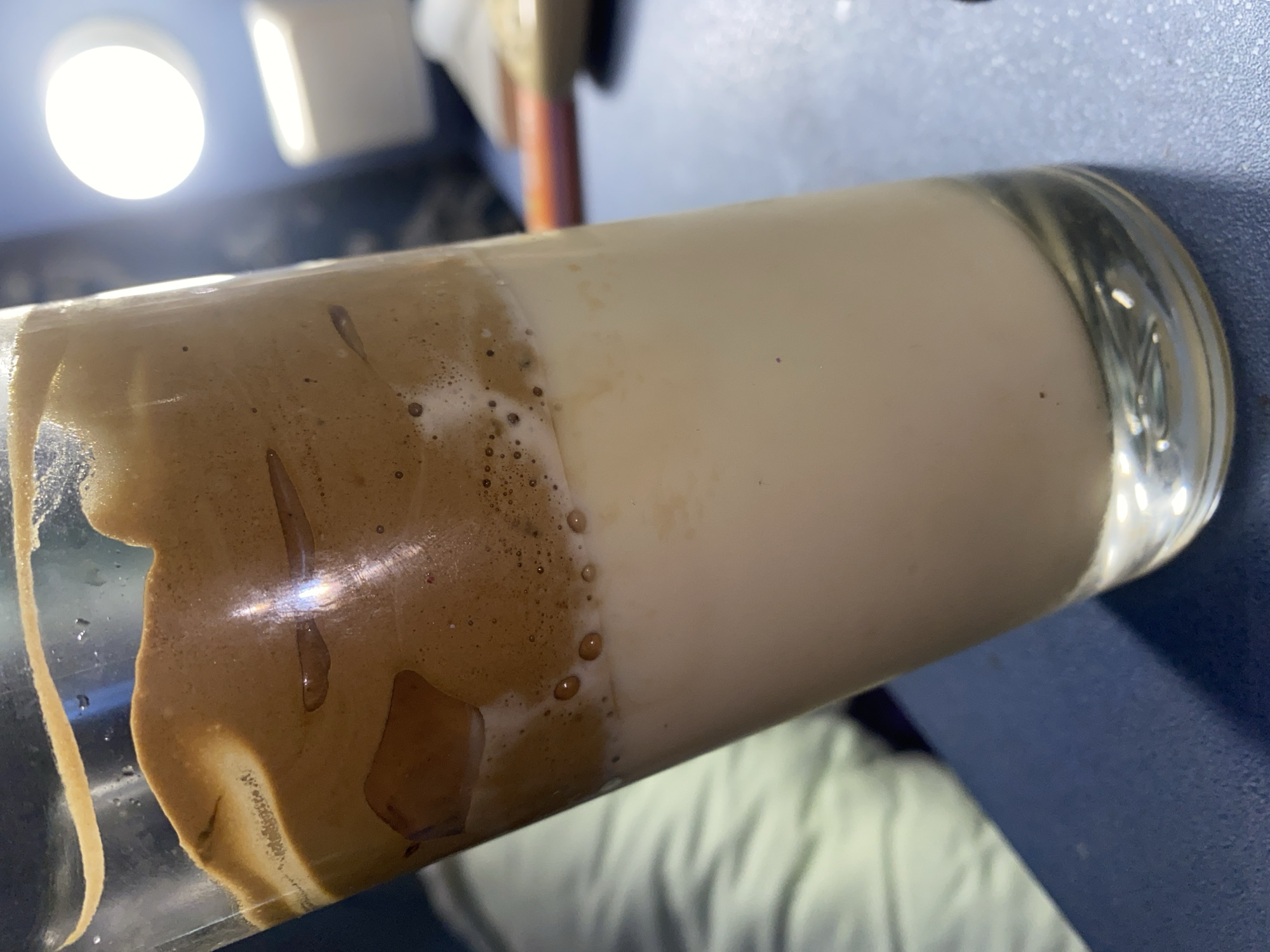A tall, clear glass filled with a layered beverage, possibly Dalgona coffee, sits on a dark blue surface. The bottom layer is a pale, milky liquid, and the top layer is a thick, foamy, brown substance with streaks of lighter brown. The glass is slightly tilted. In the background, out of focus, a bright circular light source is visible against a dark background, along with some white and orange objects. The overall lighting is dim. There are no discernible elements indicating the location of Sochi, Russia.