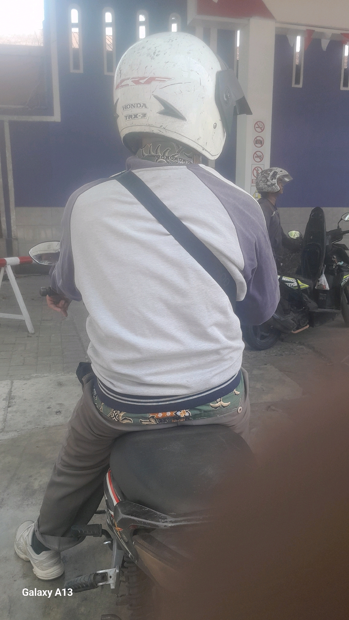 A person is sitting on a motorcycle in Tanjunggemuk, Indonesia. The person is wearing a white helmet with "HONDA TRX-3" written on it, a gray and purple long-sleeved shirt, and brown pants. The person's underwear, which has a patterned waistband, is visible above their pants. The motorcycle is black and brown. In the background, another person wearing a camouflage helmet is visible next to a motorcycle. There is a blue wall with white vertical windows and some signage behind the people. The ground is paved with concrete.