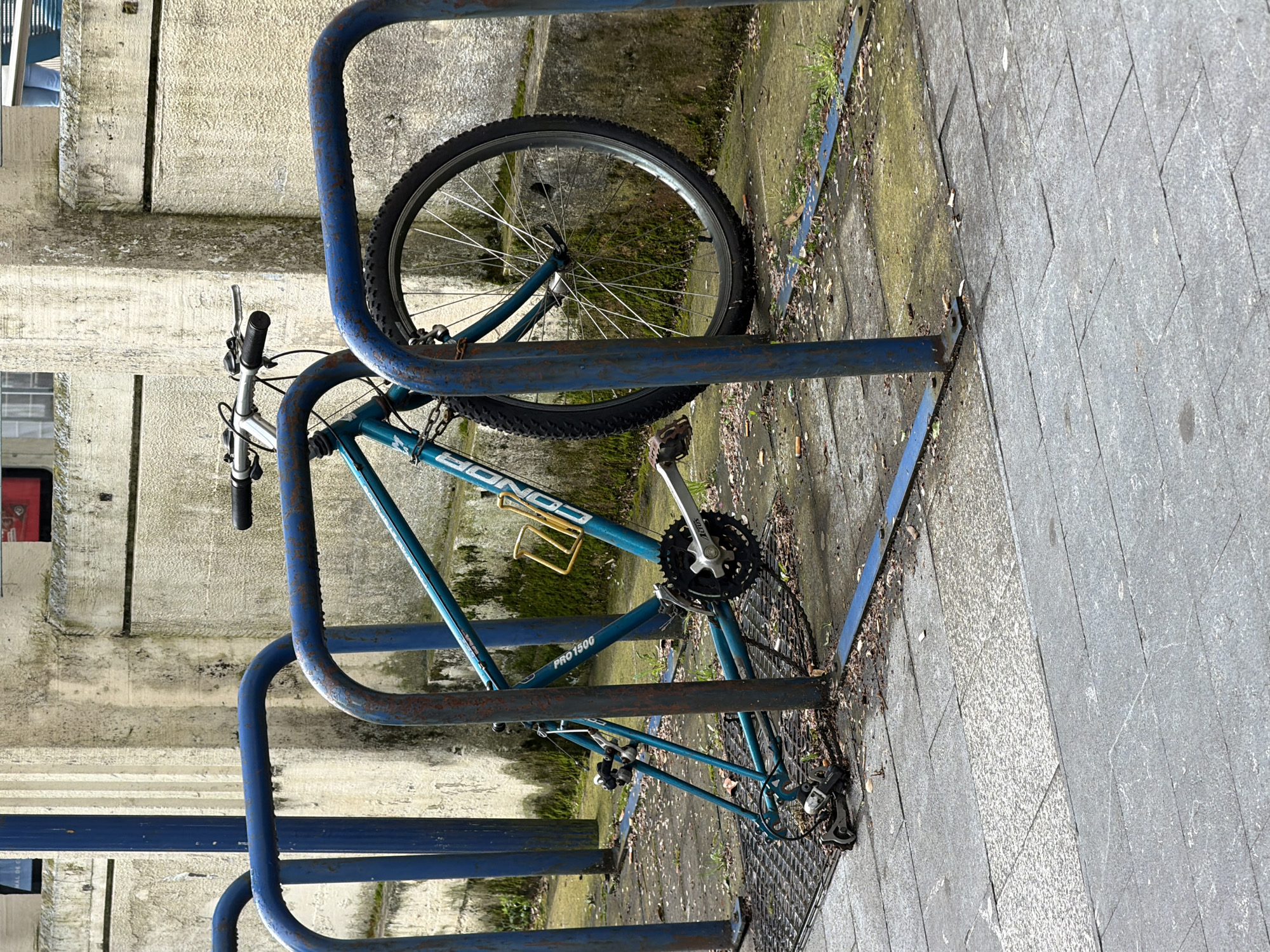 The image shows a turquoise-colored "CONOR PRO 1500" mountain bicycle locked to a blue metal bike rack in an outdoor location. The rack and bicycle appear adjacent to a light-colored, textured concrete wall and a tiled pavement. Some moss and vegetation growth are visible near the bike rack. The location appears to be an urban area in Errenteria, Spain. No people are visible in the image.
