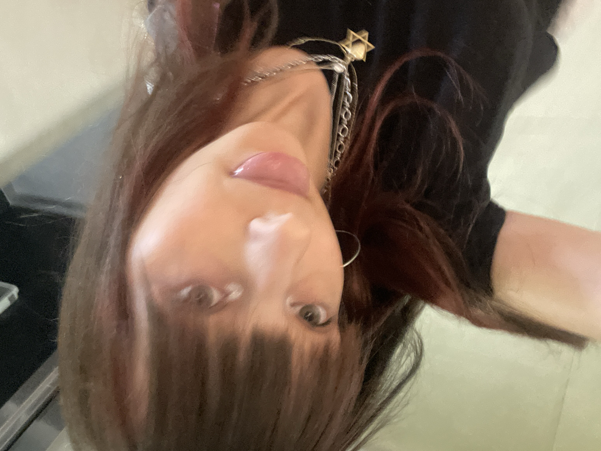 The image is a selfie taken indoors, likely in a residential setting in Raanana, Israel. The subject is a person with long, dark brown hair with auburn highlights, bangs, and fair skin. They are wearing a black t-shirt and a silver necklace with a Star of David pendant. The person is looking at the camera with their tongue slightly out. The background is out of focus and appears to be a wall or a piece of furniture, with a hint of a black object in the upper left corner. The lighting is artificial and somewhat dim.