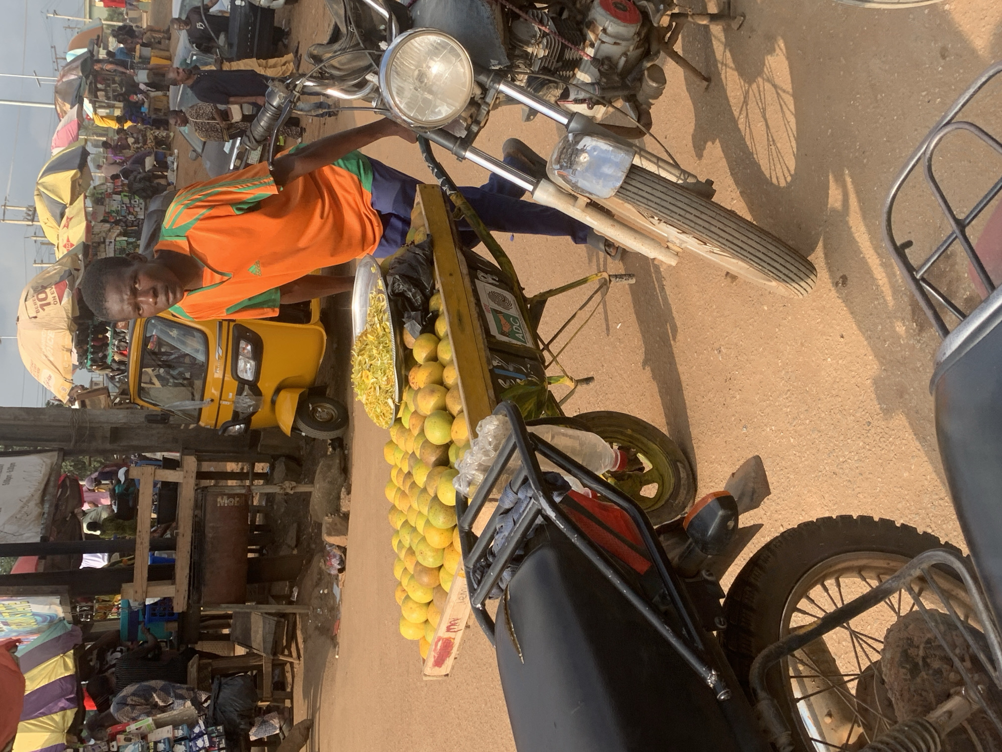 The image depicts a street scene in Shagamu, Nigeria, with a market setting. In the foreground, a man wearing an orange t-shirt with green accents and dark blue trousers stands next to a cart laden with yellow fruits, possibly oranges, and a tray of chopped yellow produce. The cart is attached to a motorcycle, which is also partially visible. Behind the man and cart, a yellow three-wheeled auto-rickshaw is parked. Further in the background, market stalls with various wares and umbrellas are visible, along with other individuals and vehicles, including what appears to be another motorcycle and a car. The ground is a sandy, dirt surface, indicative of an outdoor market. The overall impression is a bustling street market environment.