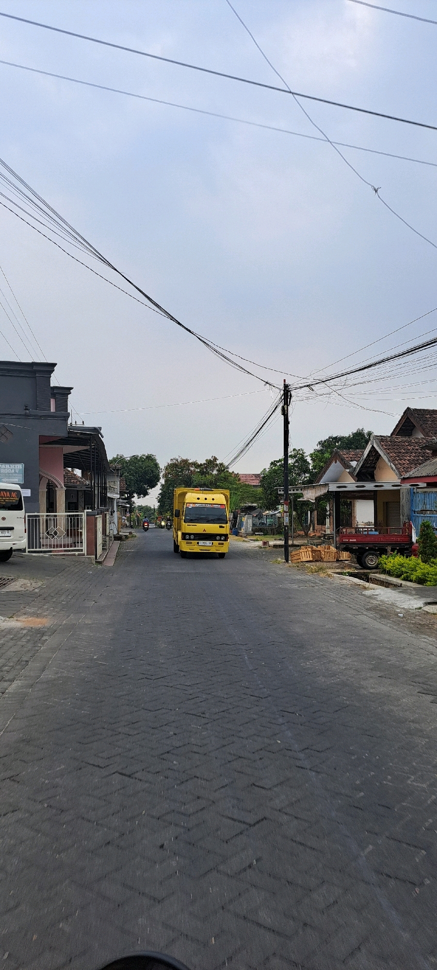 A yellow truck is driving down a paved road in Ketanireng, Indonesia. The truck is in the center of the image and is facing the camera. The road is lined with buildings and trees on either side. Overhead, a complex network of power lines crisscrosses the sky. On the left side of the road, a white van is parked in front of a building with a fence. On the right side of the road, there are more buildings and some wooden crates. The sky is overcast.