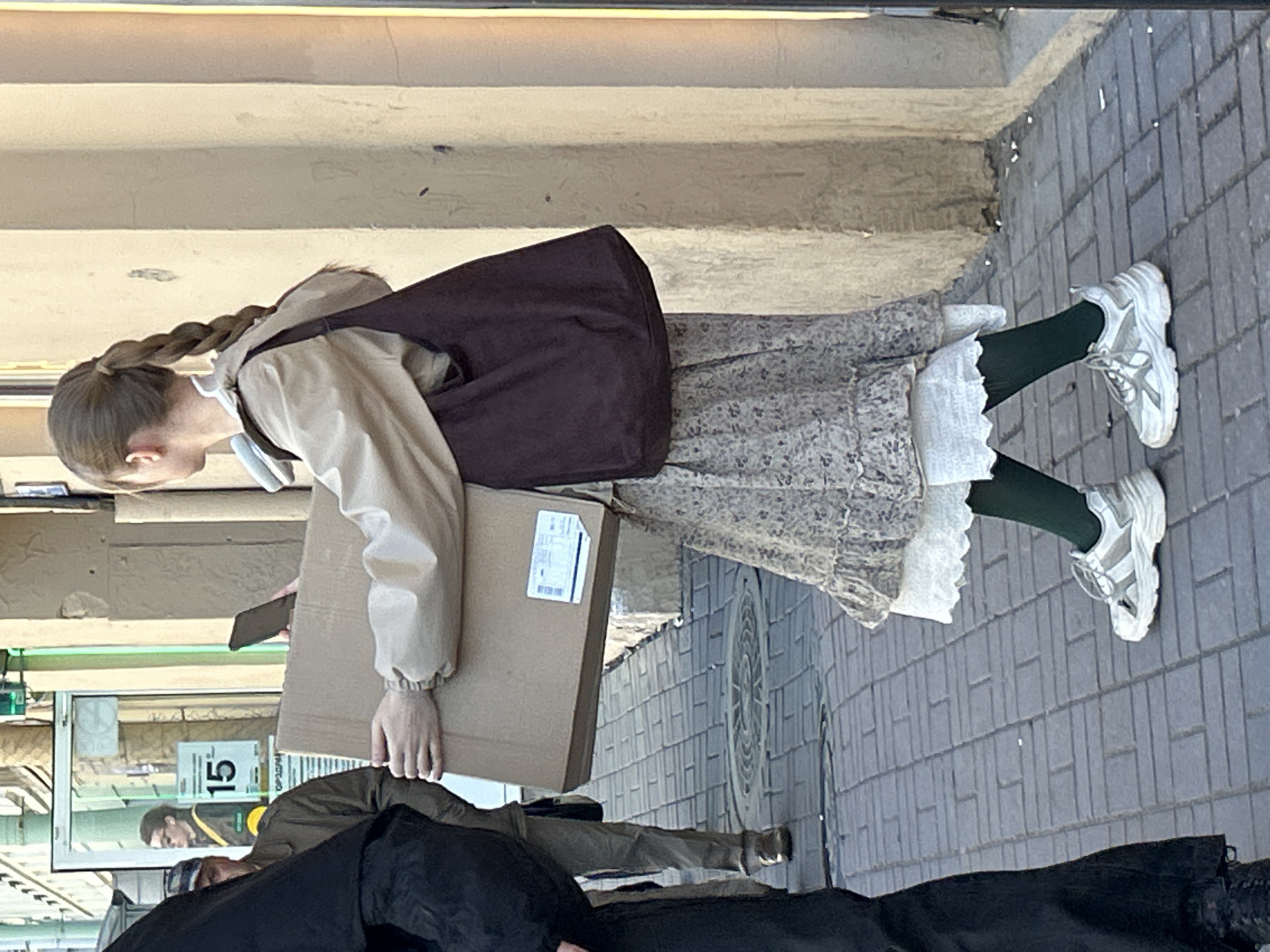 An urban street scene in Saint Petersburg, Russia, depicts a young person walking on a paved sidewalk alongside a building with shop windows. The ground is covered with grey rectangular paving stones. The main subject, appearing to be female, is seen from the side, with her head slightly bent. She has light brown hair styled in a long, thick braid. She wears a light beige or cream-colored hooded jacket, a dark brown or maroon bib-style apron or large bag over the jacket, and a long, light-colored patterned dress or skirt with a ruffled white lace hem. Dark green tights or leggings and chunky white sneakers complete her outfit. She is holding a plain cardboard box with both hands, and a smartphone is partially visible in one hand behind the box.
In the bottom right foreground, another person is partially visible, dressed in dark clothing, possibly wearing a cap. Reflected in the shop window to the left, a third person, possibly male, can be seen, wearing a dark top with a yellow stripe and looking forward. Inside the shop window, a white sign with a black border displays the number "15". The building facade above the shop window is light-colored with horizontal textured bands. The scene is brightly lit, suggesting daytime with clear weather. The overall impression is a casual, everyday street moment.