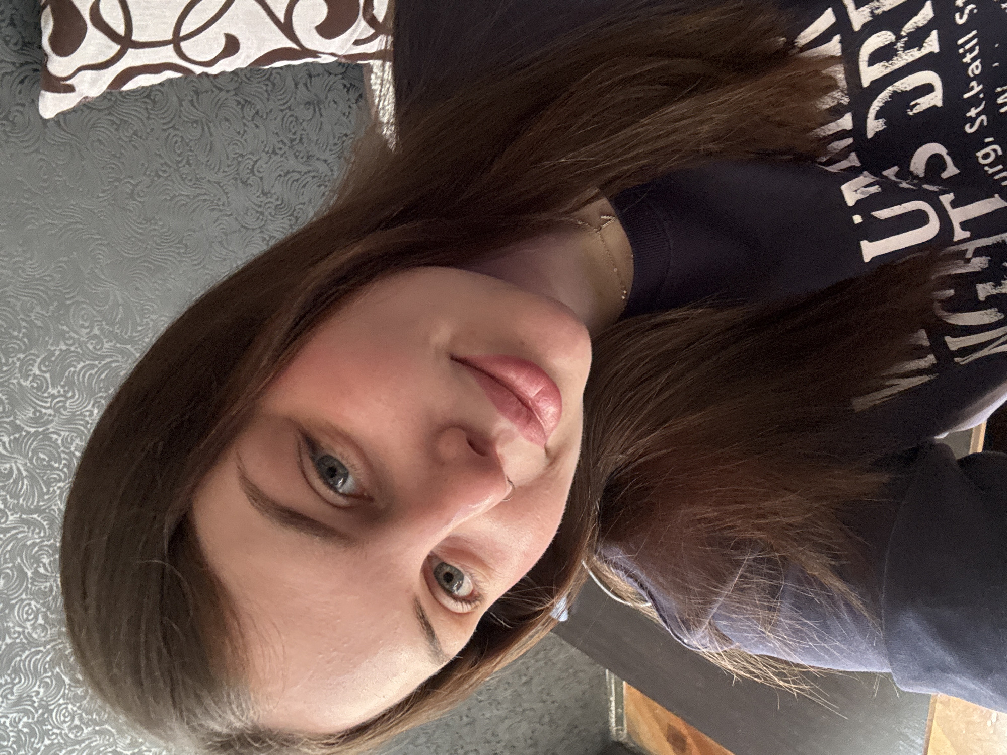 A young woman with long brown hair and blue eyes is taking a selfie. She is wearing a dark blue sweatshirt with white lettering. She has fair skin and is wearing light pink lipstick. Her head is tilted slightly to the side. Behind her, there is a patterned pillow and a gray patterned wall. The specific location in Новосельское сельское поселение, Ukraine cannot be determined from this image.