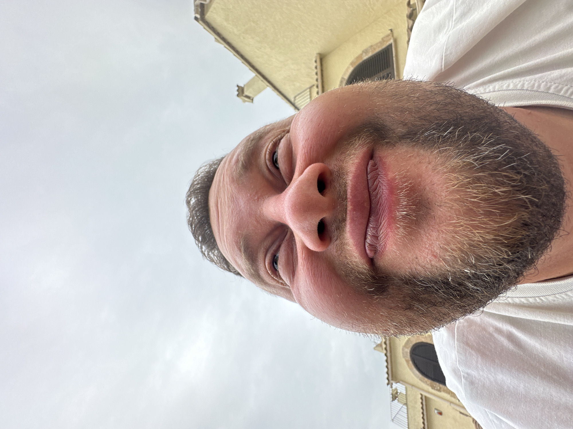 A close-up, low-angle shot captures a man's face from the chin up. He has fair skin, a light beard, and is looking upwards with his eyes partially closed and a slight smile. His cheeks are puffed out, creating a distorted perspective. In the background, a light-colored building with a terracotta-tiled roof and arched windows is visible against an overcast sky. The setting appears to be outdoors, possibly in Sant Feliu de Guíxols, Spain, given the architectural style.