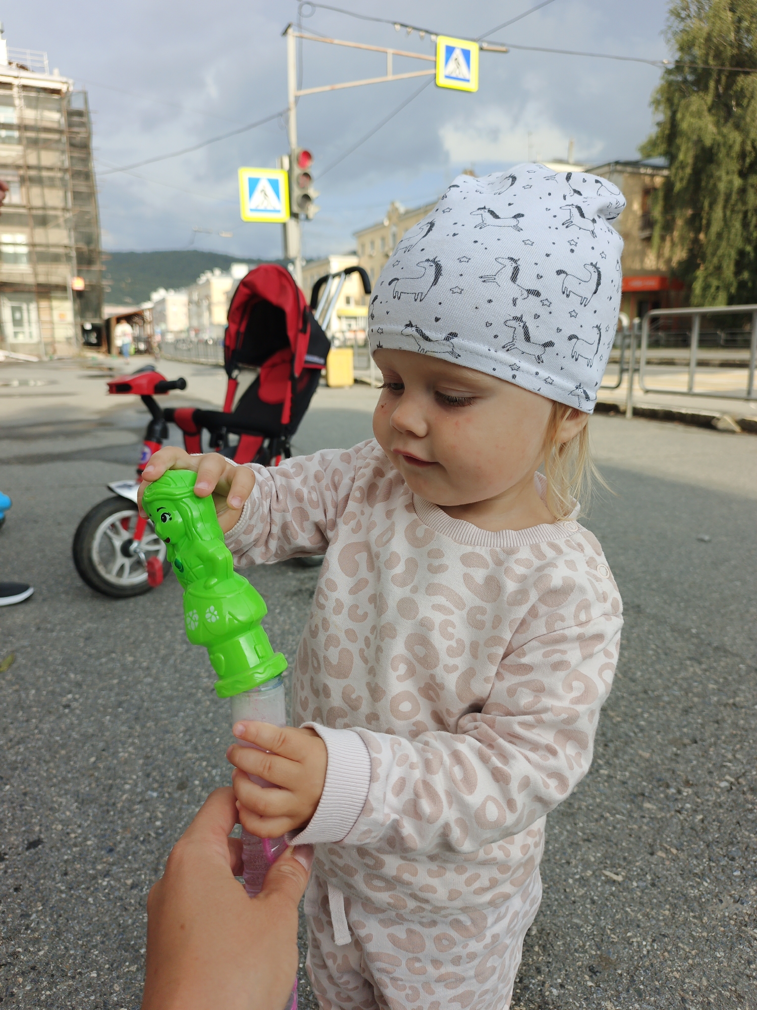 A young Caucasian child with blonde hair and fair skin, wearing a white beanie with a unicorn pattern and a light pink leopard-print tracksuit, is standing outdoors on an asphalt surface, holding a green toy in their right hand. The child's left hand is being held by an adult, whose hand is visible in the foreground and is holding a pink bubble wand. In the background, a red and black stroller is parked, and a red tricycle is also visible. A traffic light, a yellow pedestrian crossing sign, and buildings under construction are present. The sky is overcast. The location is identified as the city of Zlatoust, Russia.