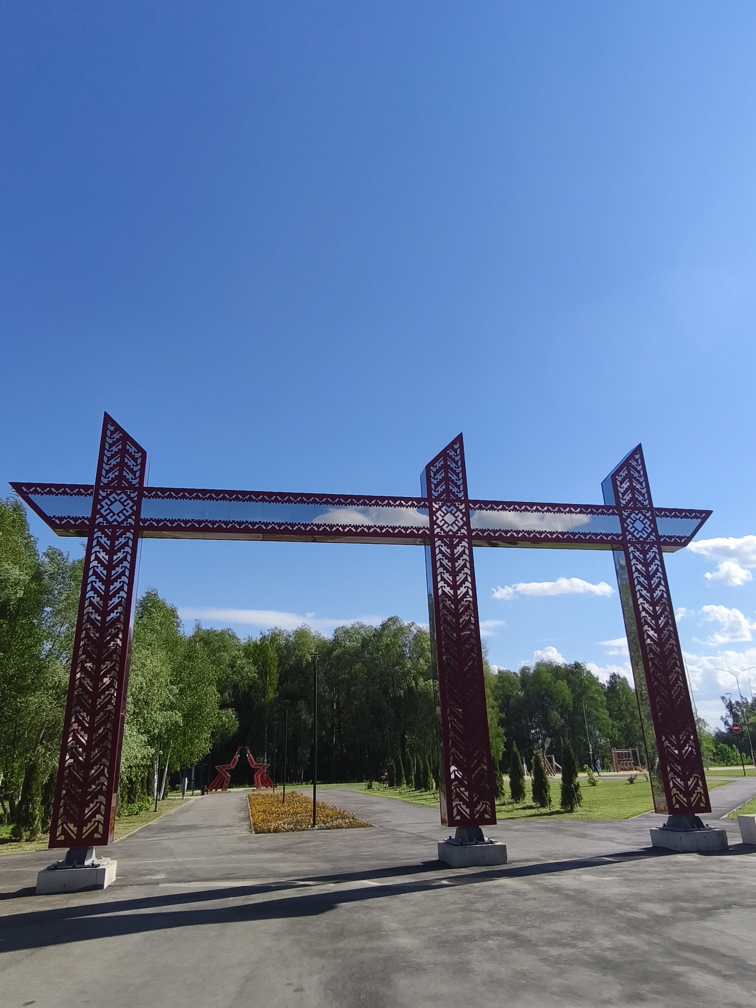 This is a ground-level, wide-angle photograph of a large, red, abstract sculpture resembling a gateway or archway, located outdoors in a park setting in Yoshkar-Ola, Russia. The sculpture is made of metal with intricate, geometric cutouts and reflective surfaces. It is positioned on a paved pathway, with a manicured lawn and a line of trees forming the background. The sky above is a clear, vibrant blue with a few scattered white clouds. In the middle distance, other red abstract sculptures are visible, along with a flower bed. The overall scene suggests a public park or recreational area, likely a point of interest or entrance to a specific zone within the park.