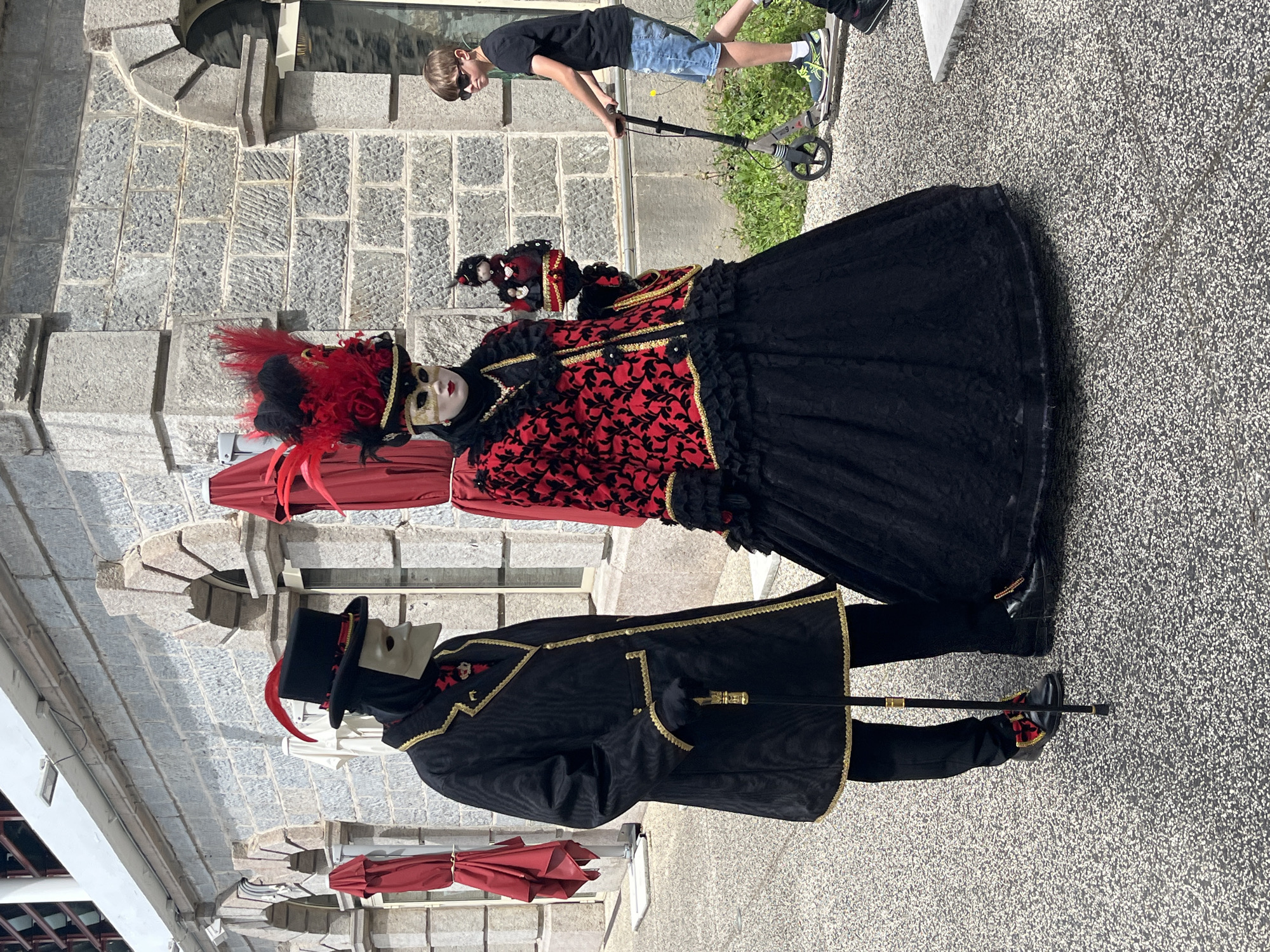 In Saint-Galmier, France, two individuals stand in ornate Venetian Carnival-style costumes. One wears a black top hat with a red feather, a black jacket with gold trim, black pants, black shoes, and a white mask. The other wears a red and black gown, black shoes, and a white mask with red feathers. Both appear to be wearing costumes related to a festival or event. A child on a scooter is in the background, near a stone wall. A gray stone pavement is visible. Parasols are visible in the background.