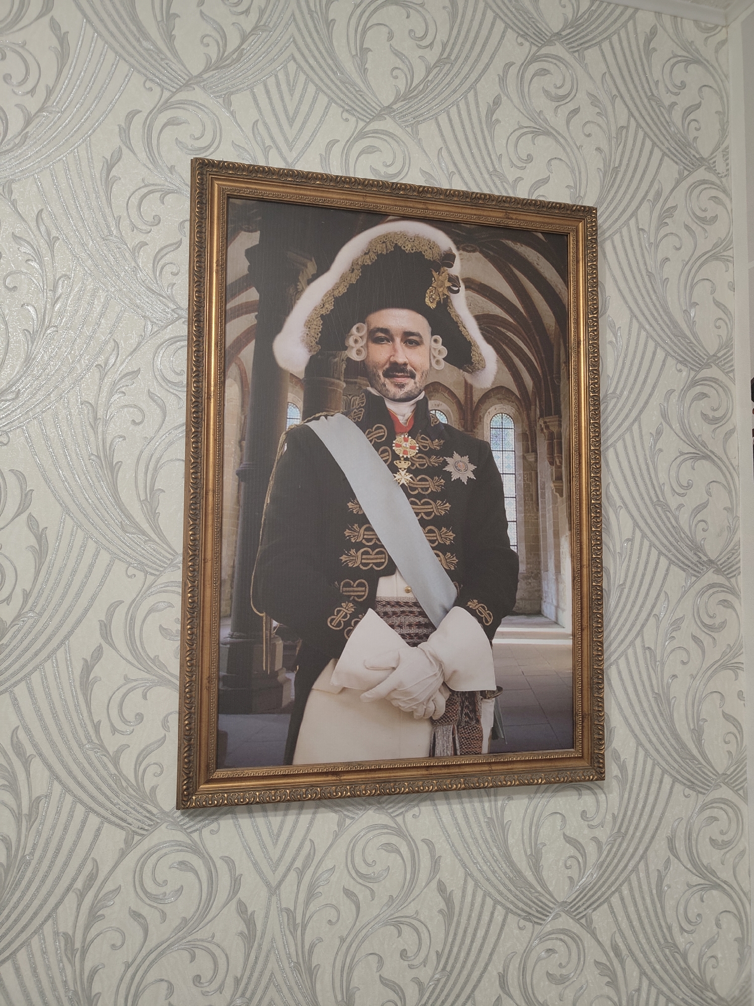 A framed portrait of a man dressed in historical military attire is hung on a wall. The man is wearing a large, feathered hat, a dark jacket adorned with gold braiding, a white sash, and white gloves. He is also wearing several medals. The background of the portrait appears to be an interior of a grand building with arched windows. The wall on which the portrait is hanging has a light-colored wallpaper with an ornate silver scroll pattern. The location is not specified as Придорожный, Russia.