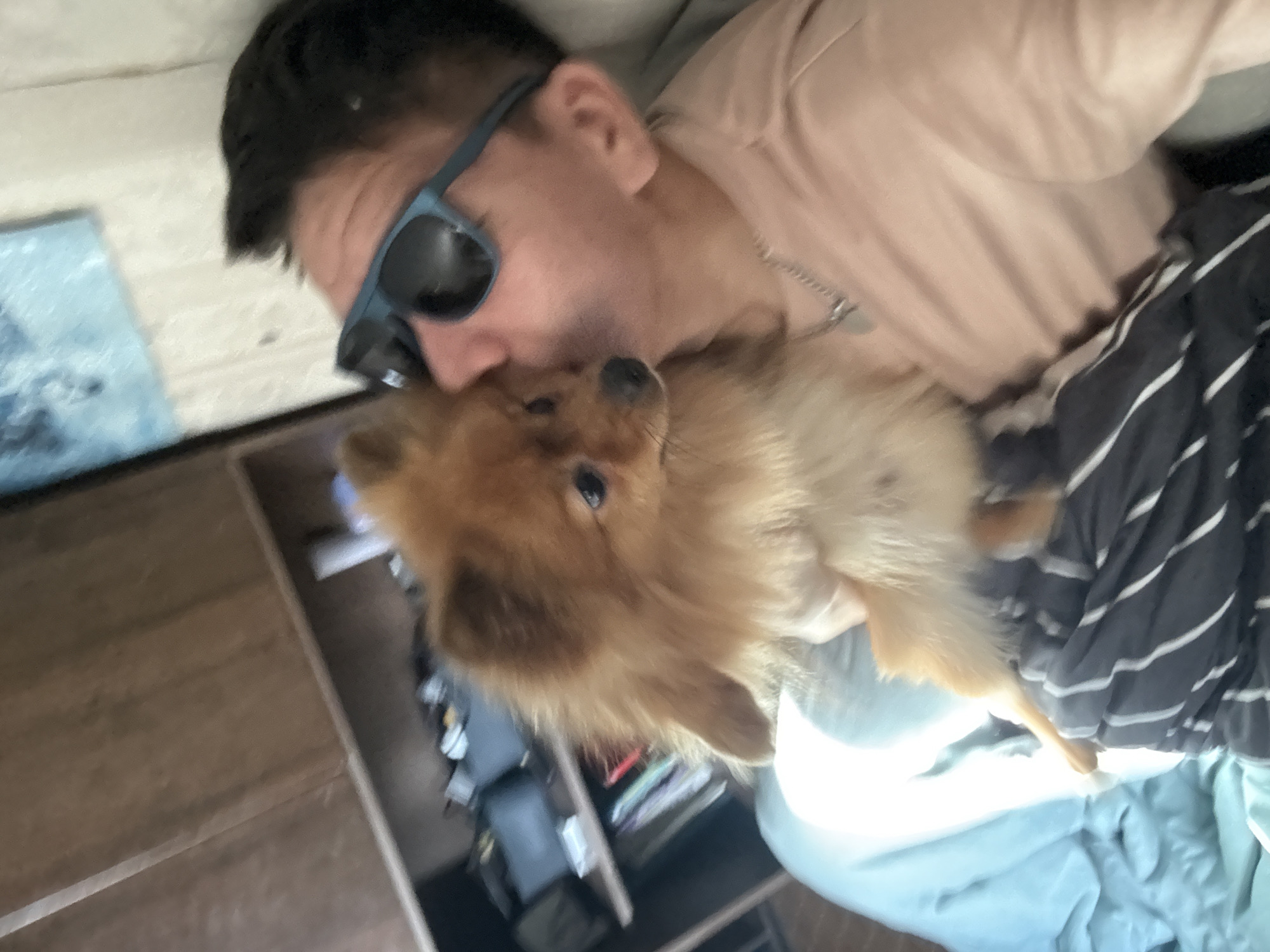 In an indoor setting, a person wearing a pink shirt and blue sunglasses holds a small, fluffy, tan Pomeranian dog, kissing its head. The person's arm is visible, along with a silver necklace. The dog has dark eyes and a dark nose. In the background, a wooden shelf unit is partially visible, containing books and various items. A light blue blanket is to the left, and a striped blanket is on the person's lap. The image appears to be a personal photograph, likely taken in a home environment. The question of the image's location in Khimki, Russia, cannot be answered from the visual information provided in the image.