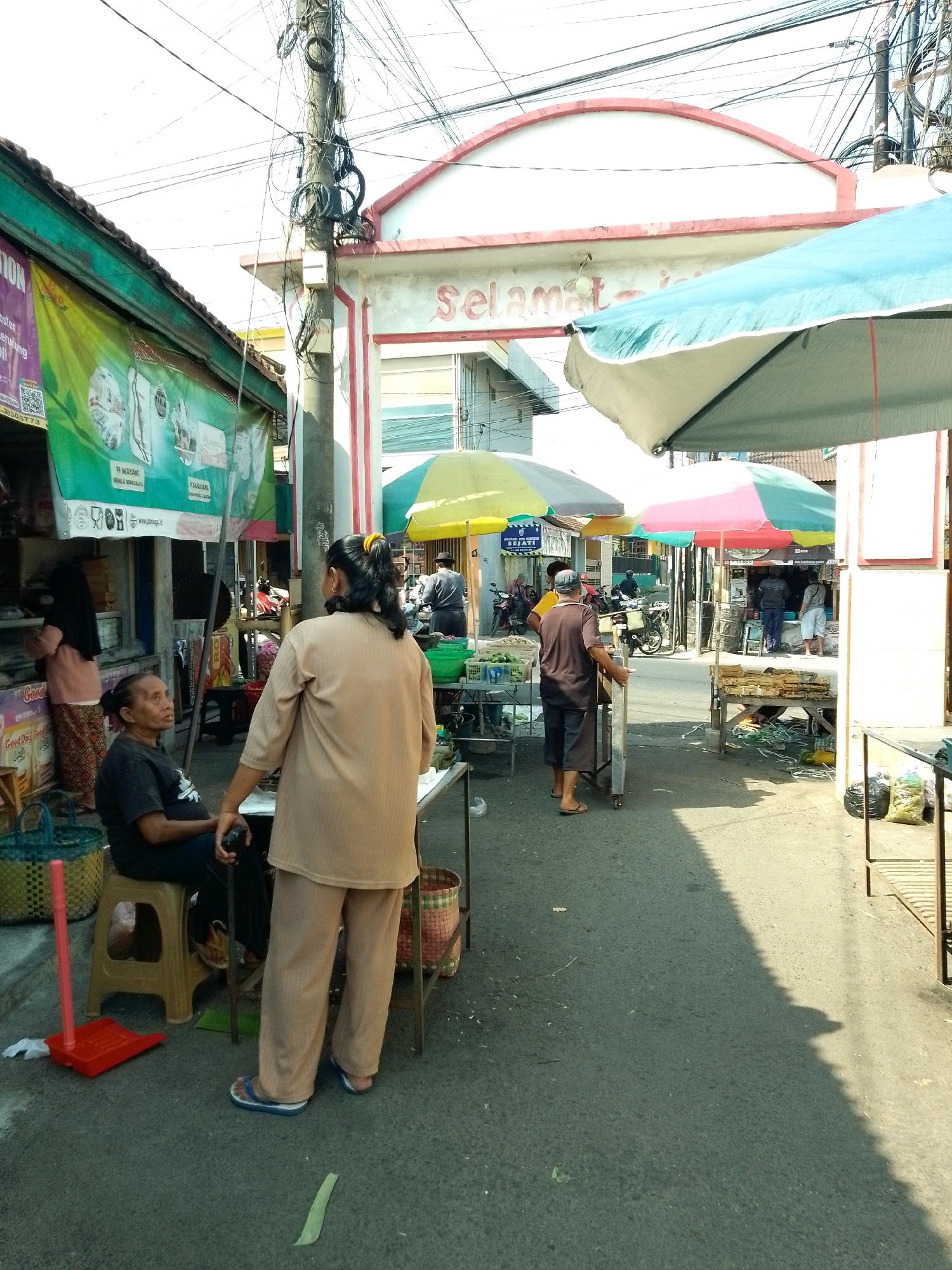 The image depicts a bustling street scene in Jember, Indonesia, likely a marketplace or a busy street lined with shops. In the foreground, a woman wearing a tan outfit and blue flip-flops stands facing away from the camera, her posture suggesting an interaction with someone off-frame. To her left, a woman in a black t-shirt and patterned skirt sits on a tan stool, looking up and to the right. A red dustpan and brush set rests on the ground near the stool.
Behind these two women, a variety of market stalls and awnings are visible. Several colorful umbrellas, some striped in red, green, and yellow, provide shade over different areas. A prominent archway with "SELAMAT DATANG" (Welcome) written in red letters spans the street in the background, indicating an entrance or a significant landmark. Various individuals can be seen in the middle ground and background, some tending to stalls, others walking or standing. Motorcycles are parked and visible in the distance, suggesting the mode of transportation common in the area. The environment appears to be an outdoor commercial space with a mix of permanent structures and temporary market setups. Power lines are strung overhead, a common sight in many urban areas. The ground is paved with asphalt, and shadows indicate it is daytime with the sun casting strong contrasts.