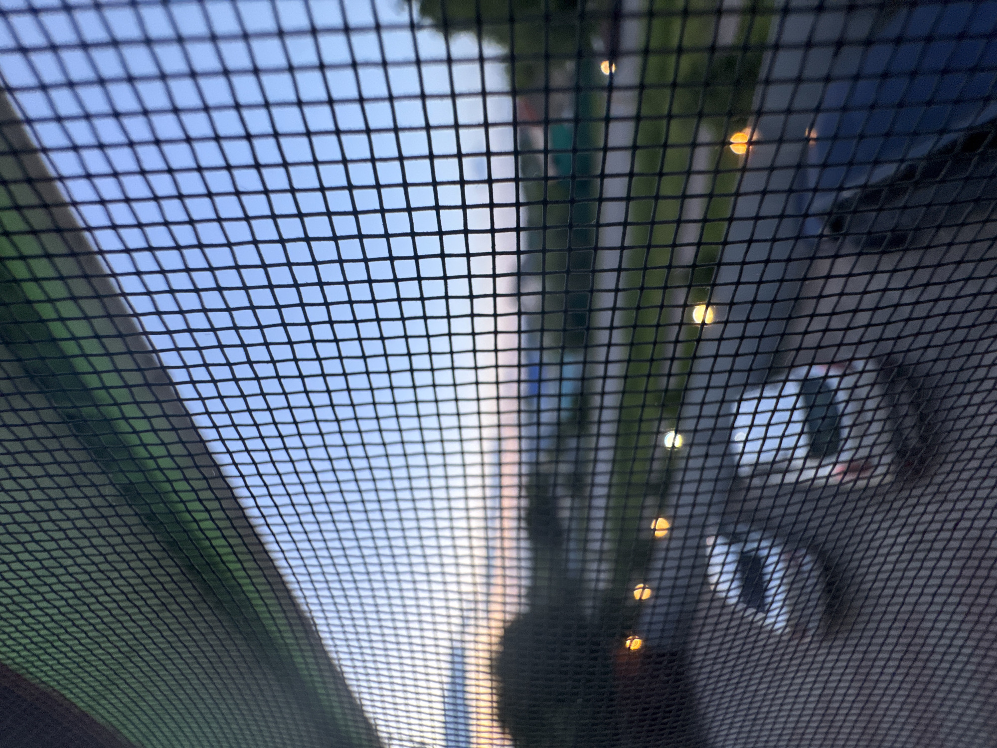 The image captures a scene viewed through a dark, fine-meshed grid or screen that covers the entire foreground. The grid pattern is distinct and evenly spaced, creating a strong visual texture that filters and blurs the background.
In the upper-left and central portions of the background, a clear, light blue sky is visible, suggesting daytime. A subtle diagonal line separates the sky from a darker, possibly green, area in the lower-left, which appears to be out of focus foliage or a distant landscape element.
The right side of the background is darker and more obscured. Several circular, yellow-orange bokeh lights are scattered throughout this section, appearing as blurred reflections or distant light sources. Through the mesh, indistinct shapes resembling parts of vehicles, such as headlights or bodywork, are vaguely discernible, along with vertical streaks that could be reflections of buildings or poles.
The perspective suggests the photograph was taken from inside an enclosure, such as a vehicle with a rear window screen or a window with a protective mesh, looking out onto a street or outdoor environment. The time of day appears to be daylight, possibly late afternoon or early evening given the visible lights. No specific activities or people are clearly visible, and there is no discernible text in the image. The scene is likely set in Mozhga, Russia, as per the provided context.