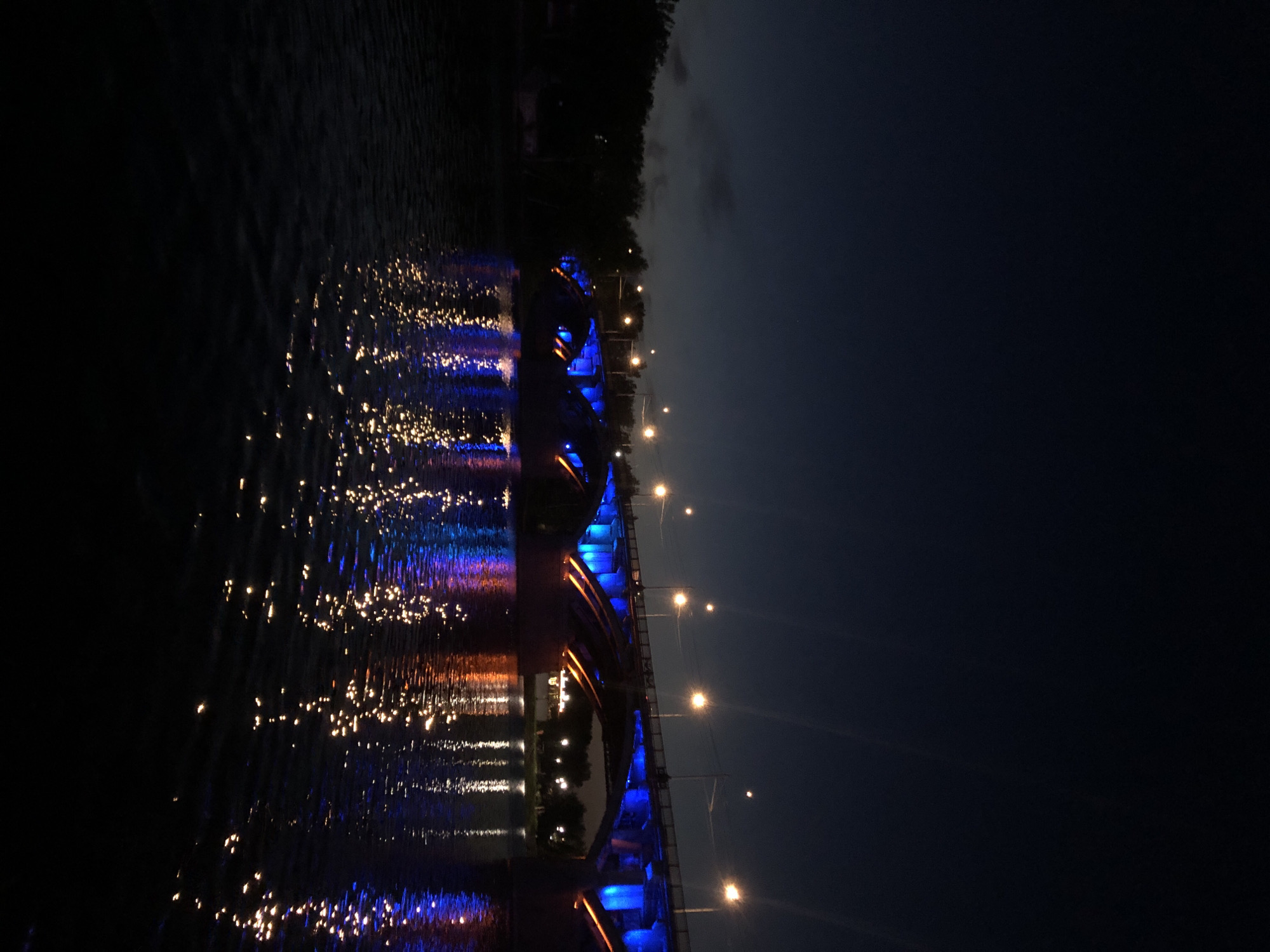 The image presents a nighttime view of the Communal Bridge (Коммунальный мост) spanning the Yenisei River in Krasnoyarsk, Russia. The sky is dark blue-black, with a subtle gradient of lighter tones near the horizon on the left. The prominent multi-arched bridge is illuminated with artificial lighting. Its primary structural elements are bathed in a vibrant blue light, while the undersides of the arches and support structures exhibit a contrasting warm orange-yellow illumination. Bright white-yellow streetlights, mounted on poles along the bridge's upper deck, cast distinct upward light streaks due to lens flare. The dark surface of the Yenisei River in the foreground and beneath the bridge reflects these lights, creating fragmented, shimmering patterns of blue, orange, and white across its rippled surface. Dark silhouettes of trees or dense foliage line the far left bank of the river, with additional faint lights discernible on the distant right bank beyond the bridge. No individuals or vehicles are visible within the frame.
