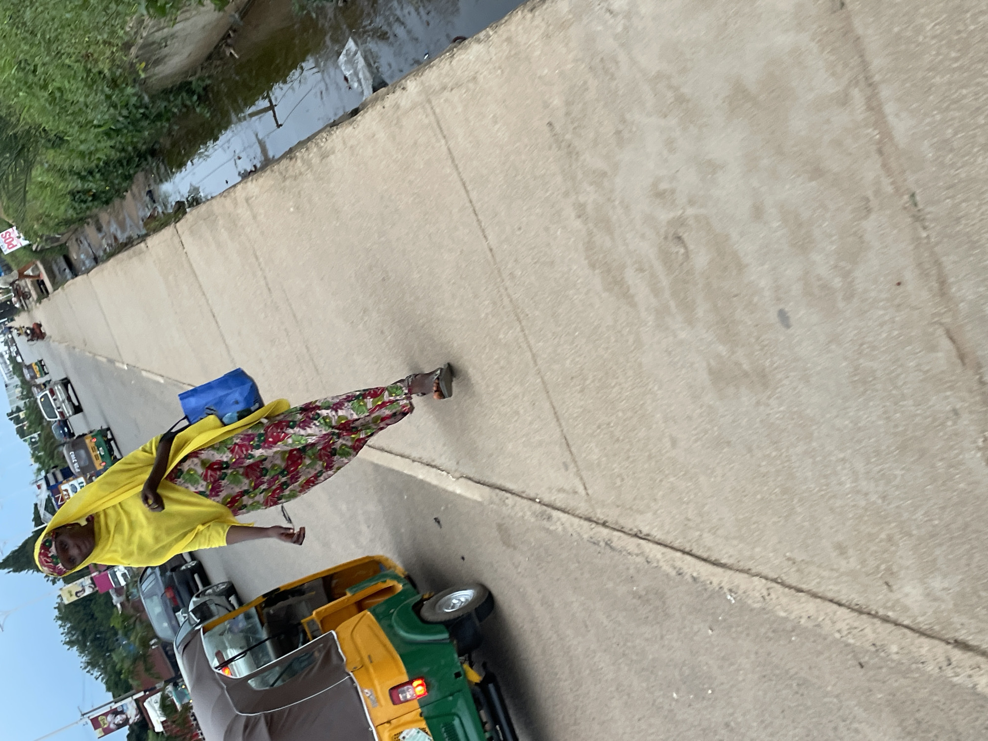 A woman in a yellow headscarf and a floral print skirt walks along a concrete sidewalk next to a drainage ditch. She carries a blue bag in her right hand. A yellow and green auto-rickshaw is parked on the street to her left. Cars and other vehicles are visible on the road in the background. The scene appears to be in an urban area.