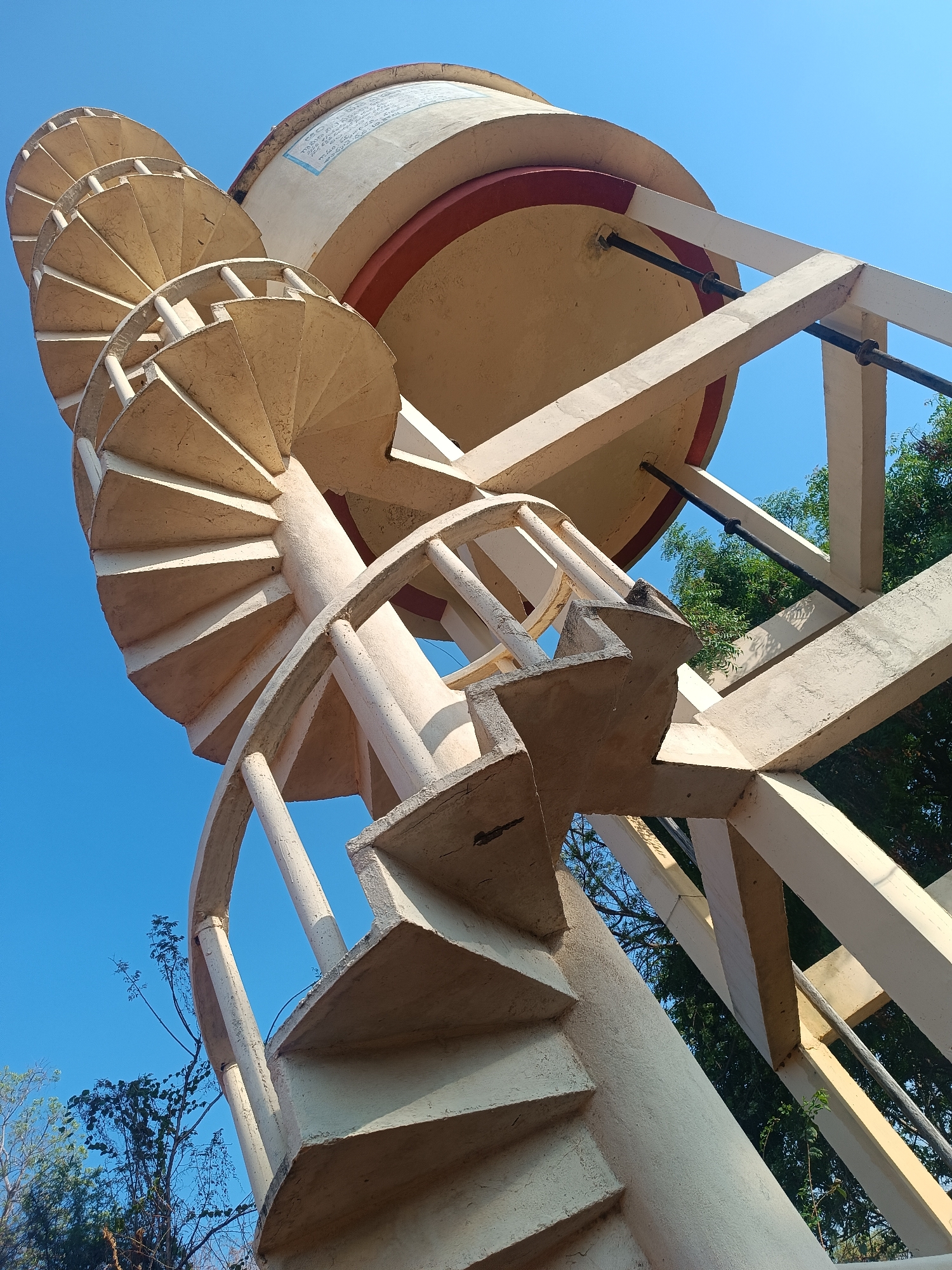 The image depicts an elevated water tank with a spiral staircase in Kammavaripalli, India. The tank is a cylindrical structure supported by a network of beams. The staircase winds around a central column, leading to the top of the tank. The sky is clear and blue. Trees are visible in the background.