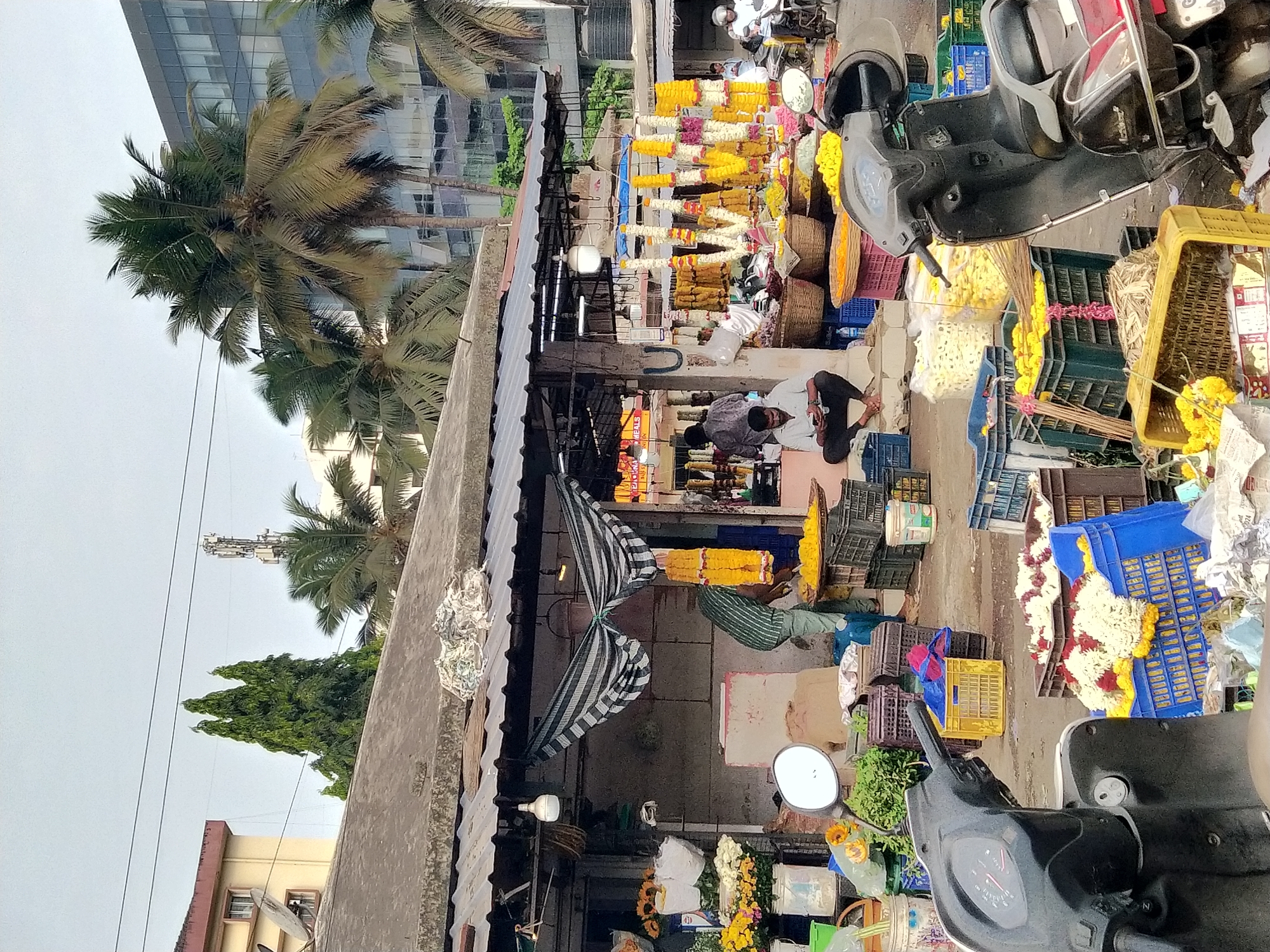 The image depicts a bustling flower market scene in Panaji, India. Two men are seated on the ground within the market area, appearing to be involved in activities related to the market's operations. A wide array of colorful flower garlands and loose flowers are displayed for sale, arranged both hanging from the stalls and in baskets/crates on the ground. Numerous plastic crates, some containing flowers, are scattered throughout the market. The scene is partially shaded by an awning supported by metal framing, with a gray and white striped cloth providing additional cover. Two scooters are parked at the edge of the market, suggesting customer transport. In the background, palm trees are visible, along with a tall modern building, contributing to the urban Indian setting.