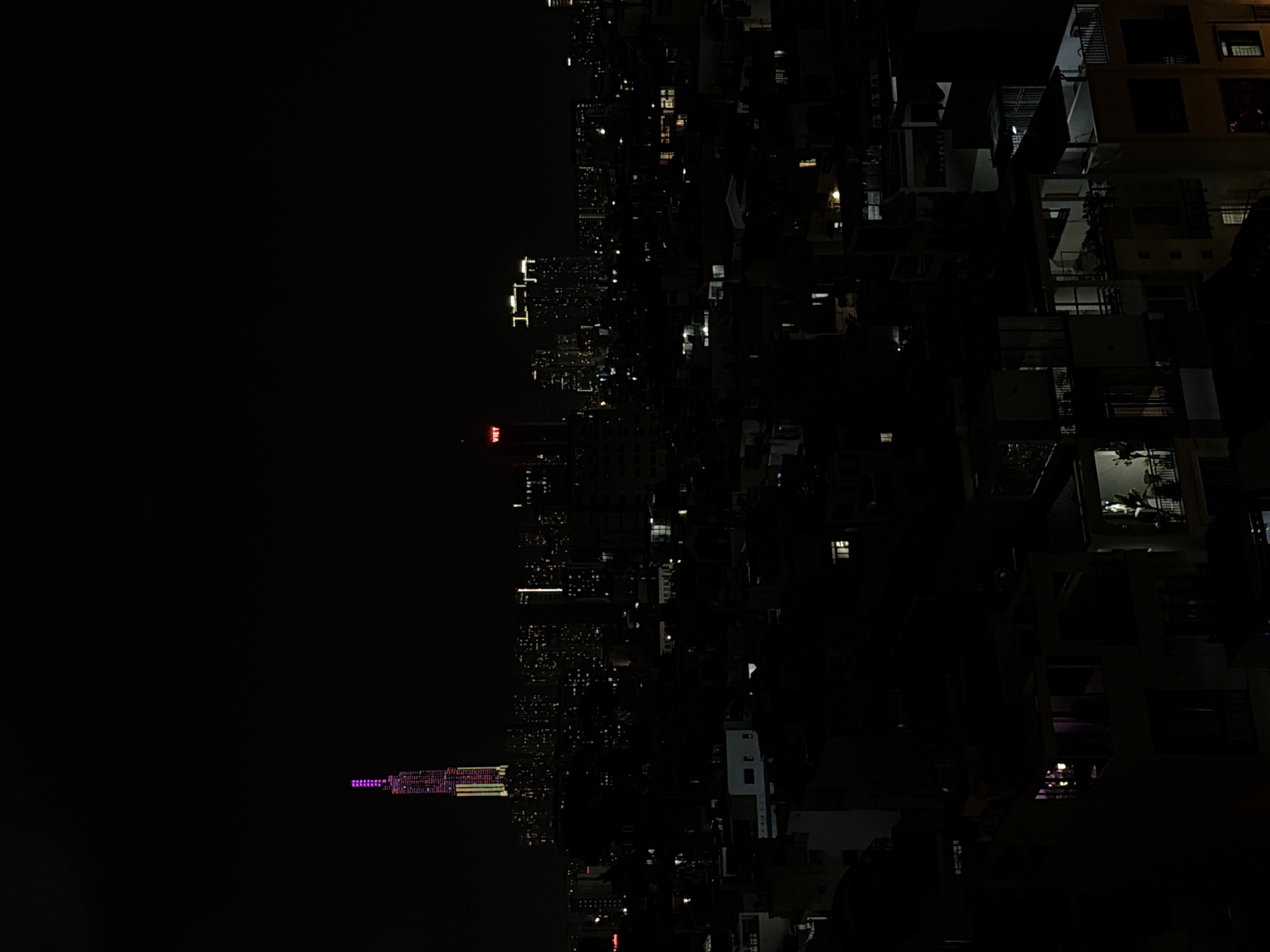 The image captures a sprawling city skyline at night, viewed from a high vantage point overlooking urban residential and commercial areas. The vast majority of the frame is dark, with the only illumination coming from countless artificial lights within buildings and structures.
In the mid-ground, a dense collection of urban buildings extends into the distance, marked by scattered windows and rooftop lights. A particularly prominent, slender skyscraper stands slightly to the left of the center, distinguished by its vibrant, complex illumination in hues of purple, pink, and other colors along its facade. Another building in the mid-ground, slightly closer and to the left, features a distinctive red illuminated sign that clearly spells out "VIET". Further back, a multitude of smaller lights twinkle across the distant cityscape, indicating a broad expanse of urban development.
In the closer foreground and to the right, larger, darker residential or commercial buildings are visible, with clearer architectural details like balconies and multiple lit windows revealing interior spaces. No individuals are discernible, and no specific activities or events are explicitly taking place; the scene is a static nocturnal cityscape.
The time of day is clearly night, evidenced by the dark sky and the sole reliance on artificial lighting. The atmosphere appears clear, with no visible fog or haze obscuring the distant lights. The image depicts a vibrant urban environment characteristic of a densely populated area, consistent with the given context of Thủ Đức, Vietnam.
