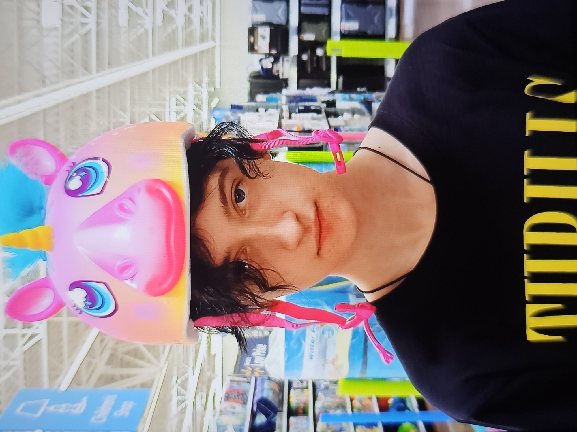A person wearing a pink unicorn helmet and a black t-shirt with yellow lettering stands in an aisle within a store. The helmet has a yellow horn and blue and pink accents. The t-shirt has large yellow block letters that appear to spell out "THRILLS". The background of the image shows shelves stocked with various products, including what appears to be bottled water and other packaged goods. Overhead lighting illuminates the store aisle. The individual's face is visible, looking directly at the camera.