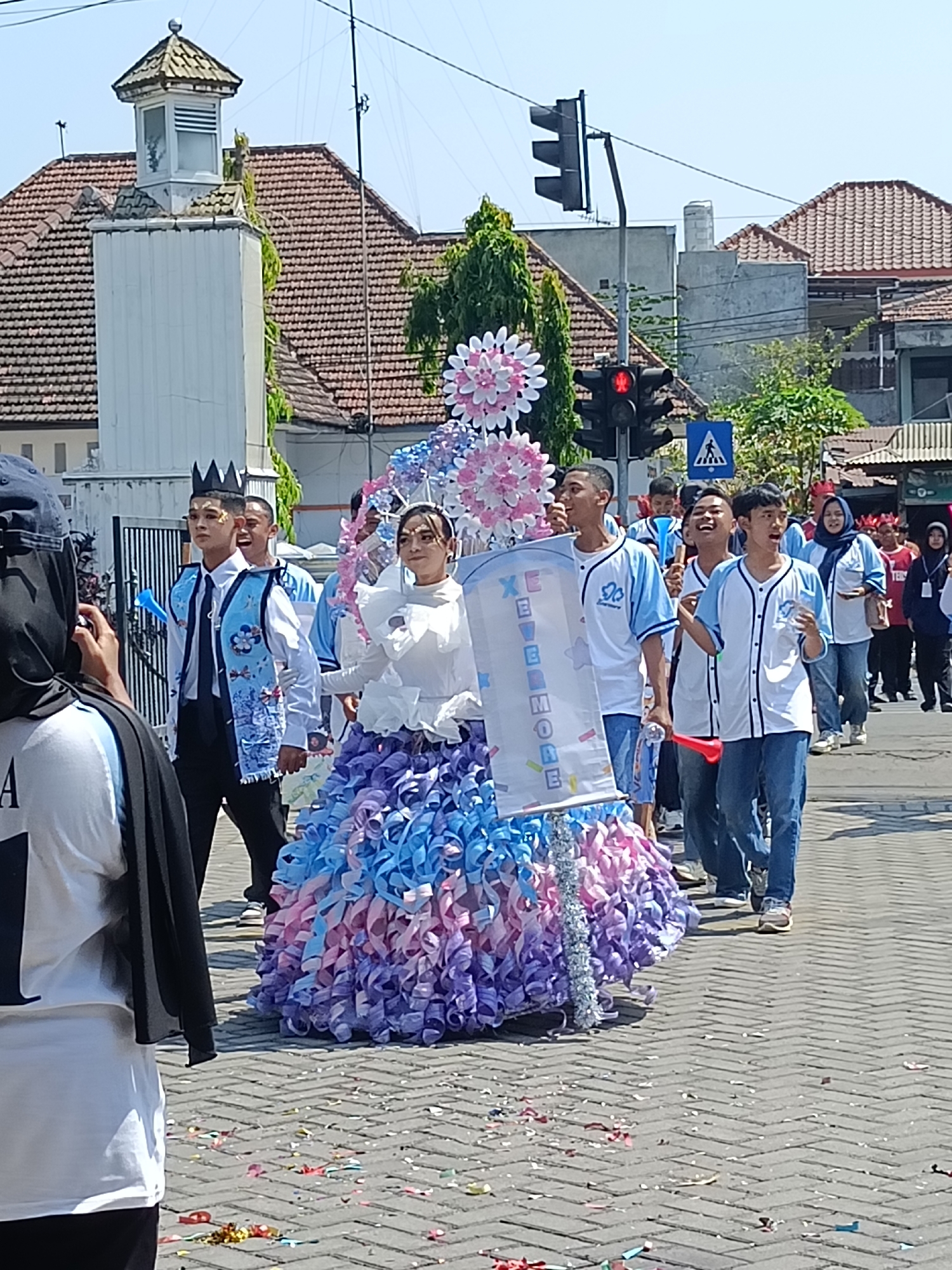 This image captures a lively procession on a street in Kediri City, Indonesia. The central figures are a group of young people dressed in festive attire. A young woman in the foreground wears a voluminous, colorful skirt made of cascading ribbons in shades of pink, blue, and purple, a white, ruffled top, and a headscarf. She is holding a large banner with lettering that appears to spell out "XEVERMORE". Beside her, a young man sports a black crown, a white shirt, a black tie, and a blue vest adorned with floral patterns. He is also wearing makeup on his face.
Behind them, a group of young men in matching light blue and white baseball-style shirts and jeans walk in the procession. Some are holding red noisemakers and appear to be cheering or singing. Other participants are visible in the background, some wearing blue shirts and jeans, and others in different attire including a woman in a red headscarf and another in a navy blue hijab. The procession is taking place on a brick-paved street, with scattered confetti on the ground.
The urban environment includes buildings with tiled roofs and visible architectural features such as a white structure with a cupola and windows on the left side of the image, and traffic lights in the background. The sky is clear and blue, suggesting a sunny day. The overall scene depicts a celebratory event, possibly a parade or festival, in Kediri City.