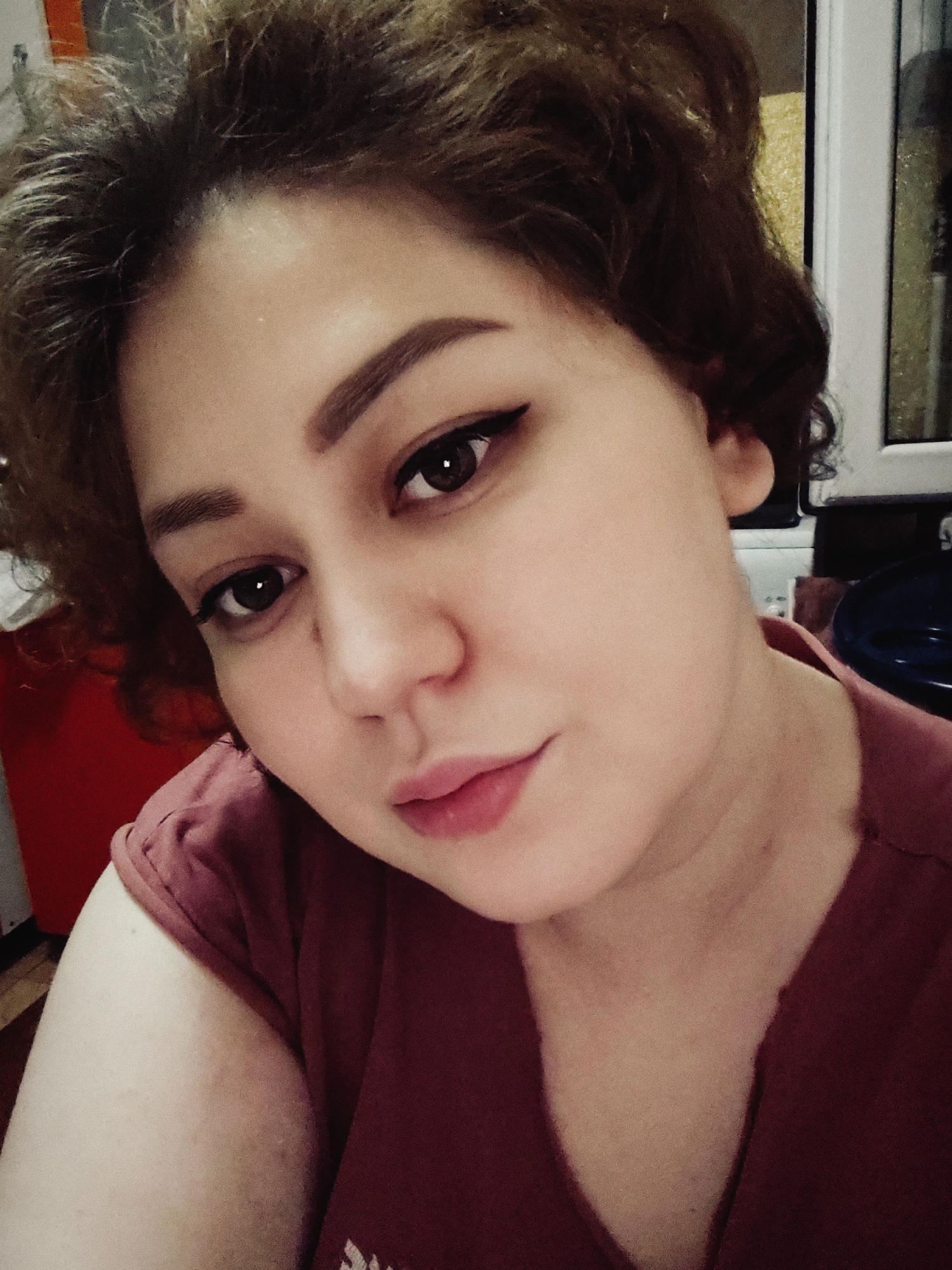The image is a close-up selfie of a young woman with light brown, curly hair and brown eyes. She is wearing a reddish-pink t-shirt. Her makeup includes eyeliner and defined eyebrows. The lighting suggests an indoor setting, possibly at night or in a dimly lit room. The background is out of focus, with hints of furniture and a window. The woman's expression is neutral with a slight hint of a smile. There are no discernible landmarks or elements that would indicate a specific location within Fergana City, Uzbekistan.
