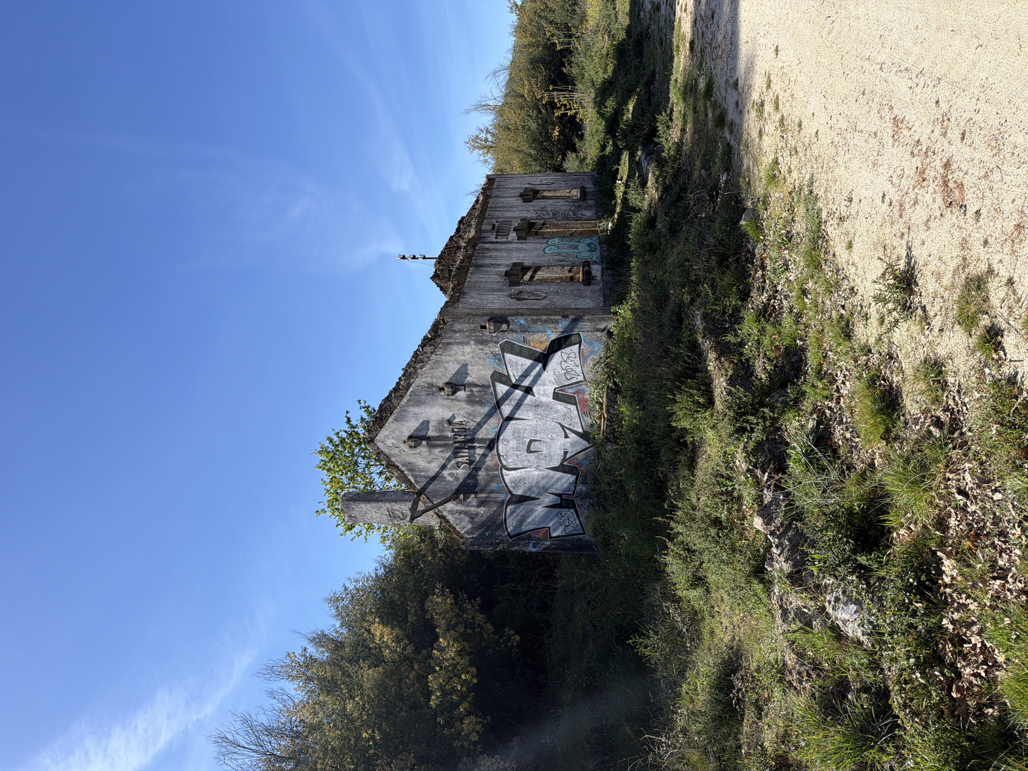 The image shows the ruins of a building in Vila Real, Portugal. The building's exterior walls are predominantly grey and covered in graffiti. The structure appears to be heavily damaged, with missing sections of the roof and walls. The building is surrounded by vegetation, with grass and bushes covering the ground. A dirt path is visible adjacent to the building. The sky is blue with light clouds. The image is taken at an angle, giving a perspective looking upwards toward the building.