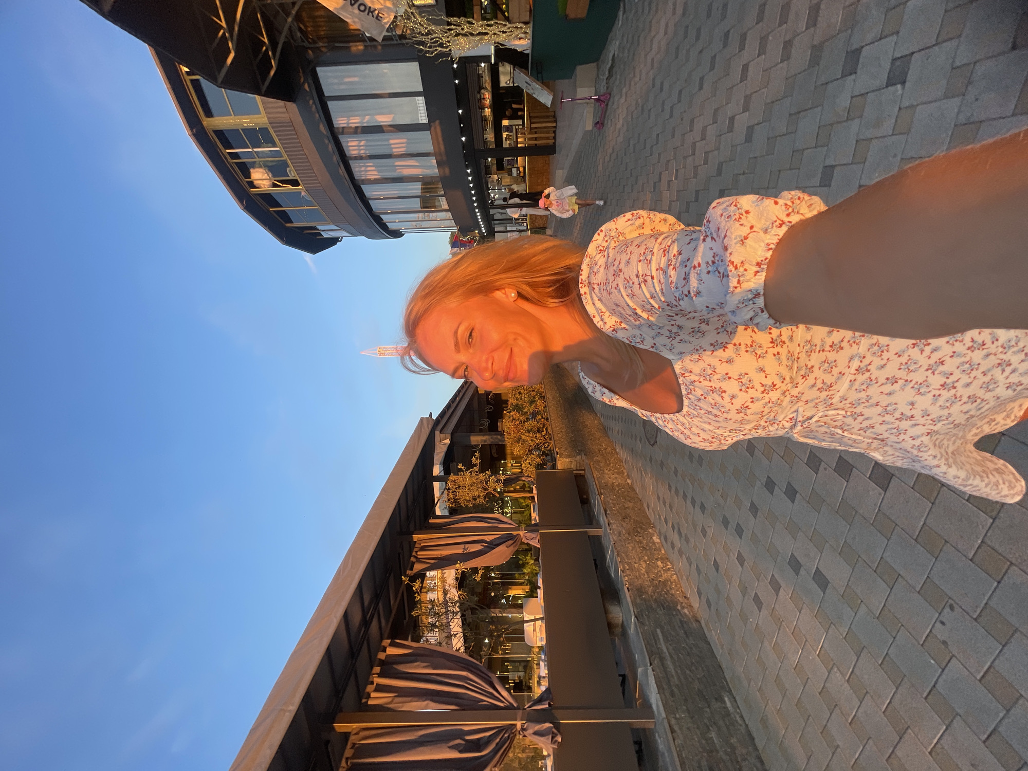 This image depicts a woman standing on a paved walkway, possibly at sunset, in Anapa, Russia. She is wearing a floral dress and holding out her arm to take a selfie. In the background, there are buildings with large windows, suggesting a commercial or restaurant area. The sky is clear and blue, and the lighting indicates late afternoon or early evening. A child can be seen in the distance running ahead. The overall scene appears to be a pleasant outdoor setting in a city.
