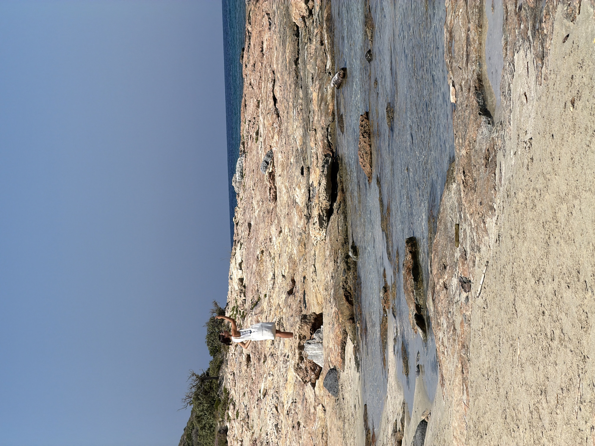 A young woman with dark hair and sunglasses, wearing a white dress and carrying a tote bag, stands on a rocky shoreline. She is holding up her phone, as if taking a picture or video. The shoreline is composed of large, flat, reddish-brown rocks interspersed with patches of sand and small tide pools. The sea is calm and blue, meeting the horizon under a clear, pale blue sky. In the background, a green, scrubby hillside rises gently. The scene appears to be a natural, undeveloped coastal area. The image is likely taken in Greece, given the characteristic rocky coast and clear blue sea, possibly in a less populated or remote area.