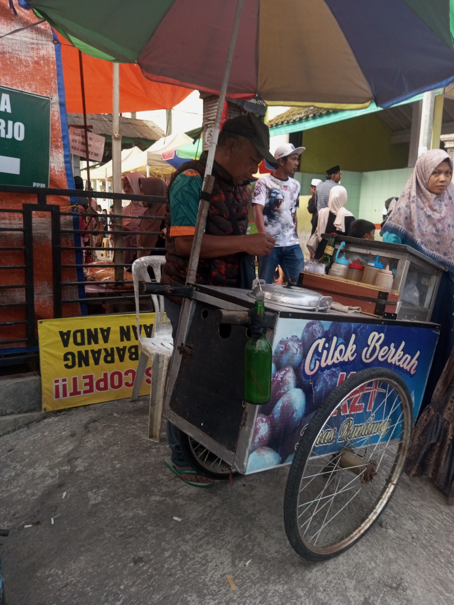 In Pandansari, Indonesia, a street scene unfolds under a colorful awning. A food cart, adorned with an image of what appears to be "Cilok Berkah" and the text "Bandung," is centrally positioned. The cart has a metal frame and a large spoked wheel on its right side. A man in a dark vest over a patterned shirt and a dark cap is seen operating the cart, his hands near its top. A green glass bottle is hanging from the cart. Behind the cart, several people are visible, including a woman in a light-colored hijab and a man wearing a white cap and a t-shirt with a lion graphic. A yellow banner with black text, reading "SCOPET!! BARANG ANDA", is placed on the ground to the left of the cart. The ground is paved and appears to be an outdoor market or street.