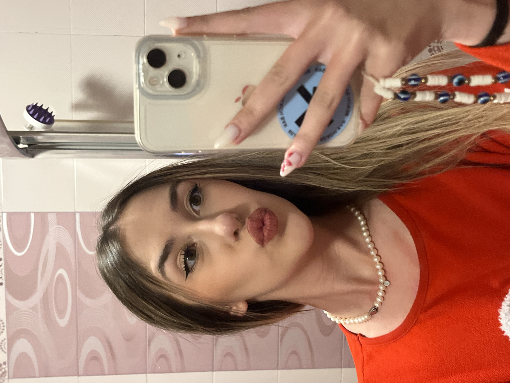 A young woman is captured in a mirror selfie, seen holding an iPhone in her left hand. She has medium-length brown hair, distinct eyeliner, and her lips are pursed in a 'kiss' or 'duck face' pose. She wears a red long-sleeved top and a delicate pearl necklace with a small heart-shaped charm around her neck. On her wrist, a bracelet adorned with blue 'evil eye' beads is visible, parts of which are intertwined with her hair. Her long, light-colored nails, possibly acrylic or gel, include red heart-shaped nail art on one finger.
The iPhone is enclosed in a clear case, showcasing its triple-lens camera system. A blue circular sticker with a prominent white 'X' is affixed to the back of the phone case, with smaller, largely unreadable text visible below the 'X', possibly "64G MOD".
The background suggests an indoor setting, likely a bathroom, characterized by light-colored tiled walls. Some tiles feature a subtle decorative pattern of pink swirls. Reflected on a shelf or ledge, a small purple scalp massager or scrubber is visible. The lighting appears to be artificial. The overall scene suggests a casual and self-expressive moment, taking place in Sofia, Bulgaria.