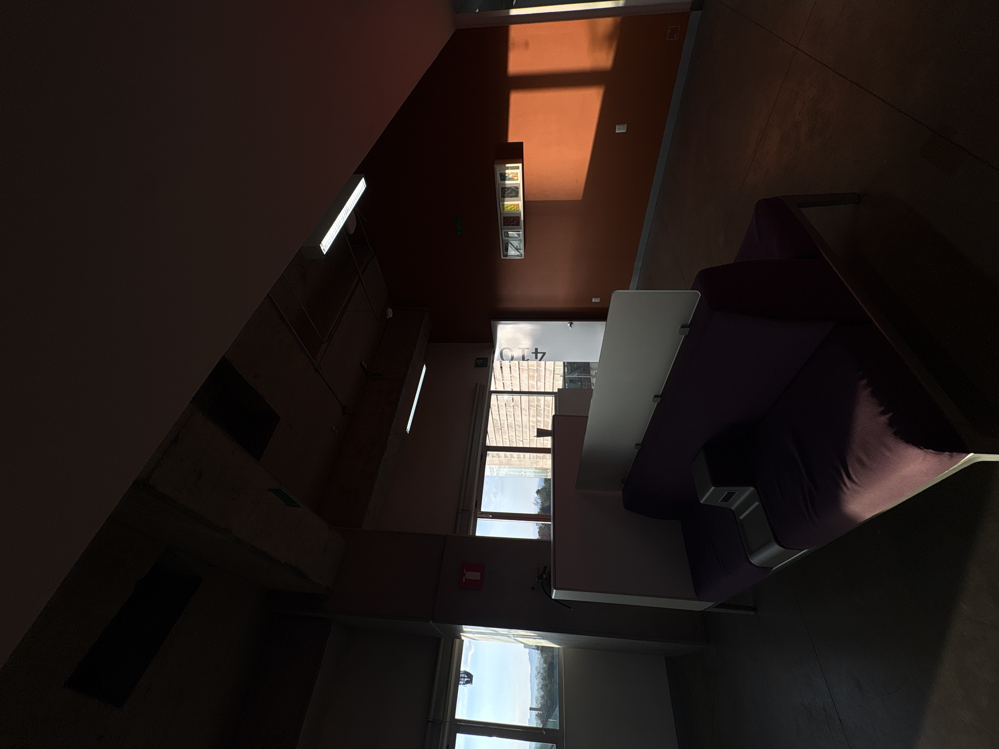 This media file presents an interior view of a modern building, captured from a high vantage point and oriented vertically. No individuals are visible, rendering the scene still and unoccupied.
The space features a prominent wall painted in a burnt orange or reddish-brown hue, which is significantly illuminated by direct sunlight streaming through a large window. This sunlight casts a distinct rectangular shadow onto the wall. Opposite this brightly lit section, a dark purple sofa or bench is positioned, accompanied by a light-colored table or counter. The floor is dark, exhibiting subtle reflections.
Artificial lighting is provided by linear fluorescent fixtures mounted on ceilings and walls; one such fixture glows brightly in the upper left portion of the image. On the orange wall, a display case or shelving unit holds several small, colorful objects.
Towards the background, multiple windows reveal an exterior view of what appears to be distant buildings or hills under a bright, clear sky, consistent with a daytime setting. Strong contrasts between sunlit areas and deep shadows characterize the overall lighting. A red sign, indicating a fire extinguisher, is mounted on a concrete wall adjacent to one of the windows. Above a doorway, a sign clearly displays the text "G 4.7". The architectural style, linear lighting, and functional signage suggest a commercial or institutional building environment, possibly a lounge area or hallway.