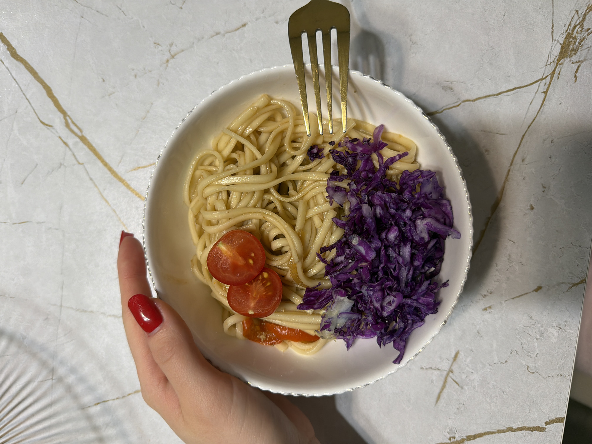 An overhead shot displays a person's hand holding a white bowl filled with a meal, set against a light-colored, possibly marble or quartz, countertop with prominent golden veining. The hand, positioned on the lower left, features nails painted with bright red polish, gently supporting the bowl.
The bowl contains three main components: a generous portion of light-colored pasta, likely linguine or a similar flat noodle, occupying the left and central parts of the bowl. On the lower left, two halves of bright red cherry tomatoes are nestled within the pasta. To the right, a vibrant pile of shredded purple cabbage adds a striking contrast in color and texture. A golden-colored fork is inserted into the pasta and cabbage on the upper right side, indicating the meal is ready for consumption.
The white bowl has a decorative, scalloped edge, possibly with a subtle metallic rim. The background surface's distinct golden streaks across its white base provide an elegant and clean setting. The lighting is even and appears to be artificial indoor lighting. No text is visible in the image. This scene is set in Chaikovsky, Russia.