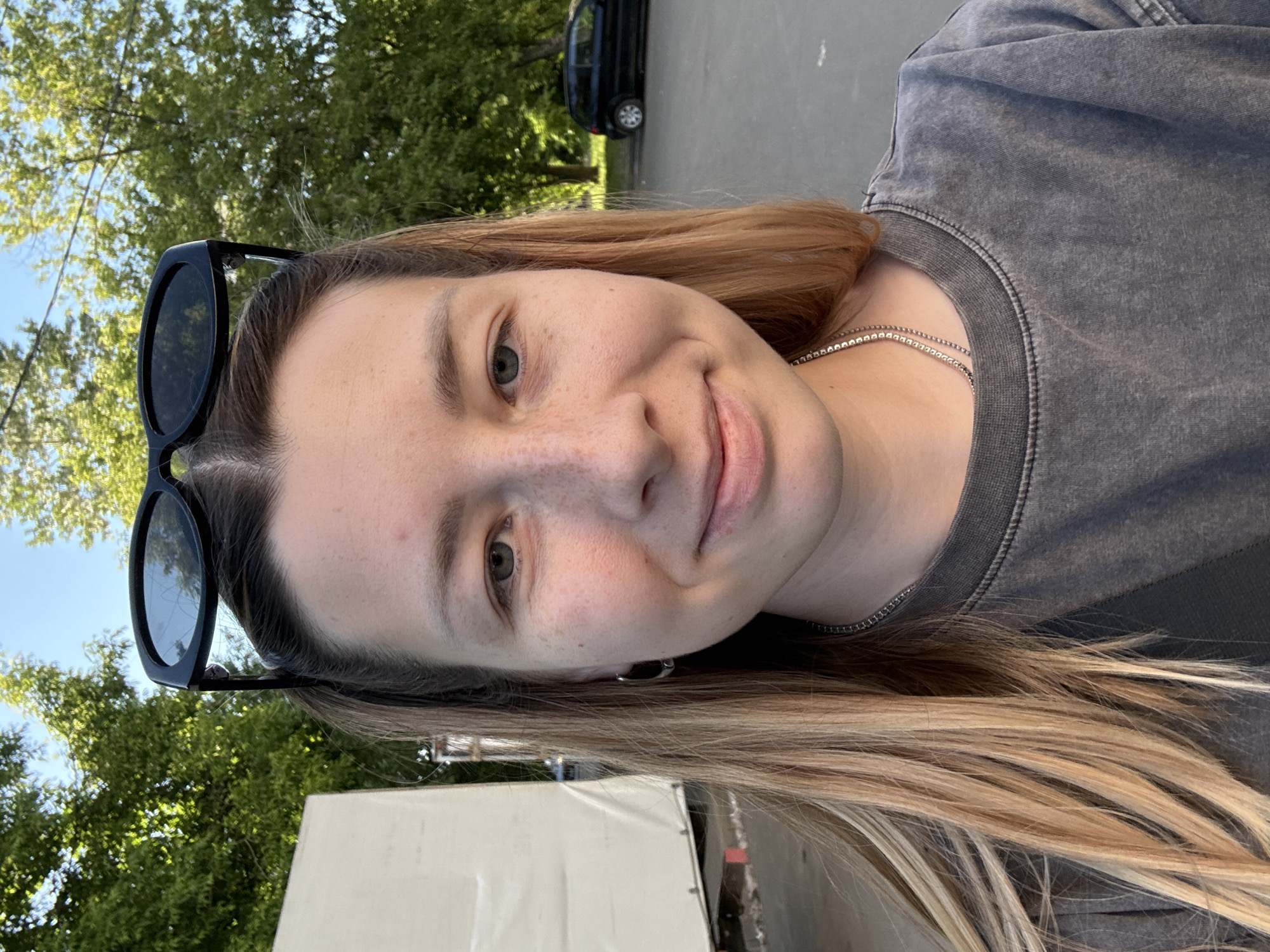 The image displays a frontal self-portrait of a young adult female positioned centrally outdoors under clear daylight. She has long, light brown hair with blonde highlights, styled back with black sunglasses perched on her head. Her complexion is fair with visible freckles across her nose and cheeks. She has light-colored eyes and a subtle closed-mouth smile directed towards the camera. A small silver hoop earring is visible in her left ear, and a thin silver chain necklace hangs around her neck. She is wearing a charcoal or dark grey, textured, long-sleeved crew-neck top.
The background shows an urban or suburban outdoor environment. To the subject's left, a large, off-white or light-colored canvas-like structure, possibly a tarp or the side of a vehicle or tent, is partially visible. To her right and extending into the distance, green foliage from trees is prominent against a clear blue sky. A paved surface, resembling a street or parking lot, occupies the lower right background, where a black compact car is parked in the mid-distance. The lighting indicates a bright, sunny day in a temperate climate.
While specific landmarks are not discernible to pinpoint an exact address, the general urban green space and street environment are consistent with many public areas within the city of Moscow, Russia.