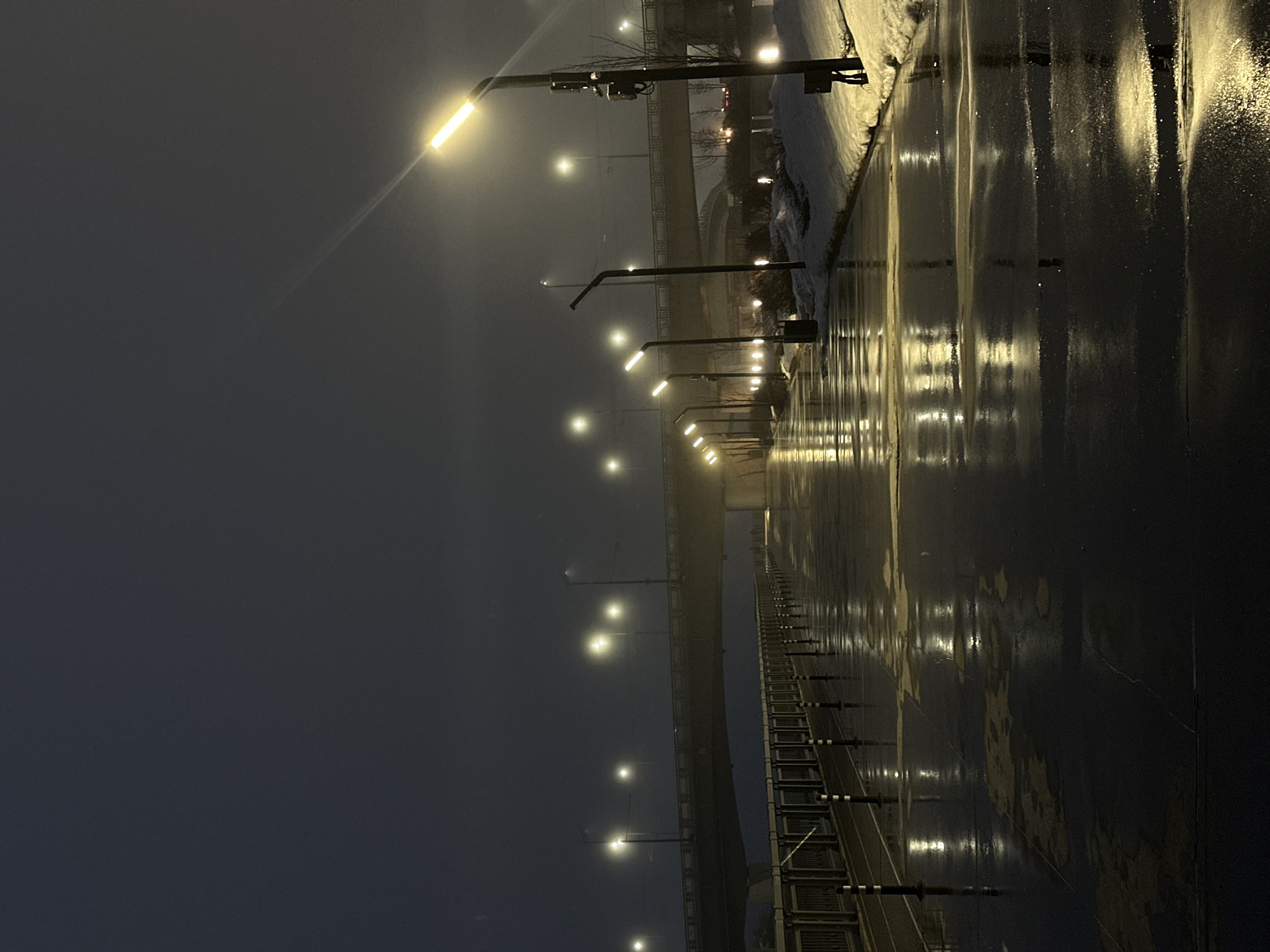 The image depicts a wet, dark street illuminated by a row of streetlights in Kazan, Russia. The street reflects the light from the lamps, creating streaks of light on the asphalt. A barrier runs along one side of the street. There appears to be snow on the edge of the road. A bridge structure is visible in the background. The scene is dusky, with a heavy sky. The image is rotated, so the scene is viewed at an angle.