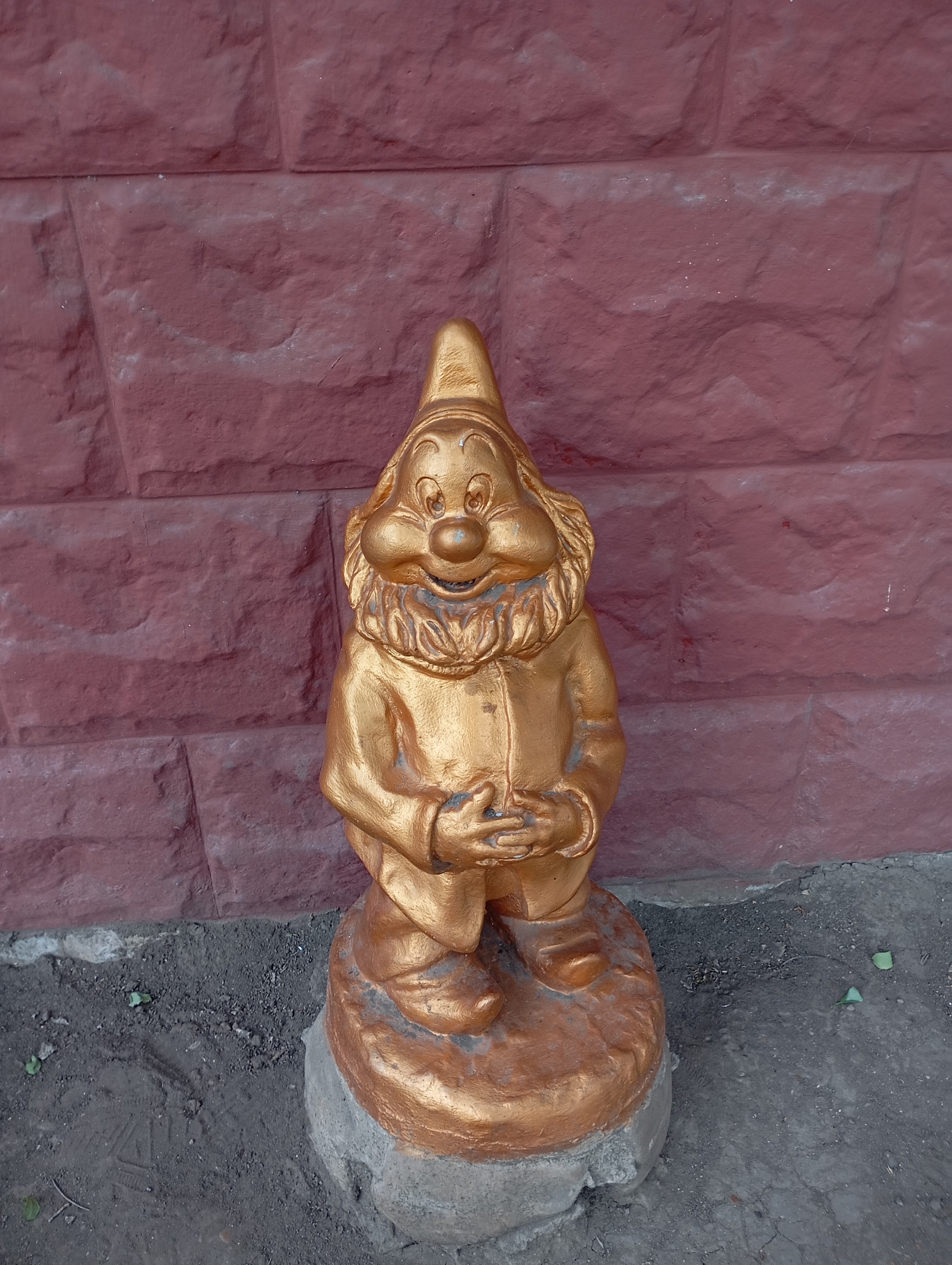 A gold-painted garden gnome statue stands in front of a textured, reddish-brown brick wall. The gnome is depicted with a smiling face, a beard, and is wearing a pointed hat and a coat. It is standing on a circular base, which is itself placed on a patch of dirt and gravel. The wall behind the gnome has a rough, uneven surface, with visible grout lines between the bricks. The ground in front of the gnome is a mix of dark soil and small stones, with a few green leaves scattered around. The lighting suggests it is daytime, with no harsh shadows visible. The overall scene appears to be outdoors, likely in a garden or yard setting. There is no indication of specific location within Tiraspol, Moldova.