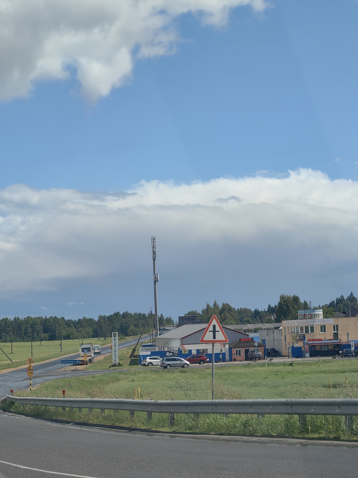 The image depicts a roadside scene in the vicinity of Фаніпальскі сельскі Савет, Belarus. A two-lane asphalt road with a metal guardrail in the foreground curves to the left. On the right side of the road, a grassy area is bordered by a metal fence. Beyond the fence, a commercial area is visible with several buildings. One building has a sign that reads "АВТО - КУЗОВНОЙ РЕМОНТ," indicating automotive body repair services. Another building has a sign with Cyrillic lettering that is partially obscured. A red triangular road sign with a black cross symbol is positioned on a pole, indicating a crossroads.
To the left of the road, a rural landscape unfolds with a field and a line of trees in the distance. A traffic sign with the number "70" is visible on the left side of the road, likely indicating a speed limit. Further down the road, a white truck with a trailer is driving away from the camera. A cell tower stands tall in the background, to the right of the trees. The sky is partly cloudy with a mix of blue sky and white and gray clouds. The overall scene suggests a functional but somewhat utilitarian commercial and road environment in a semi-rural Belarusian setting.
